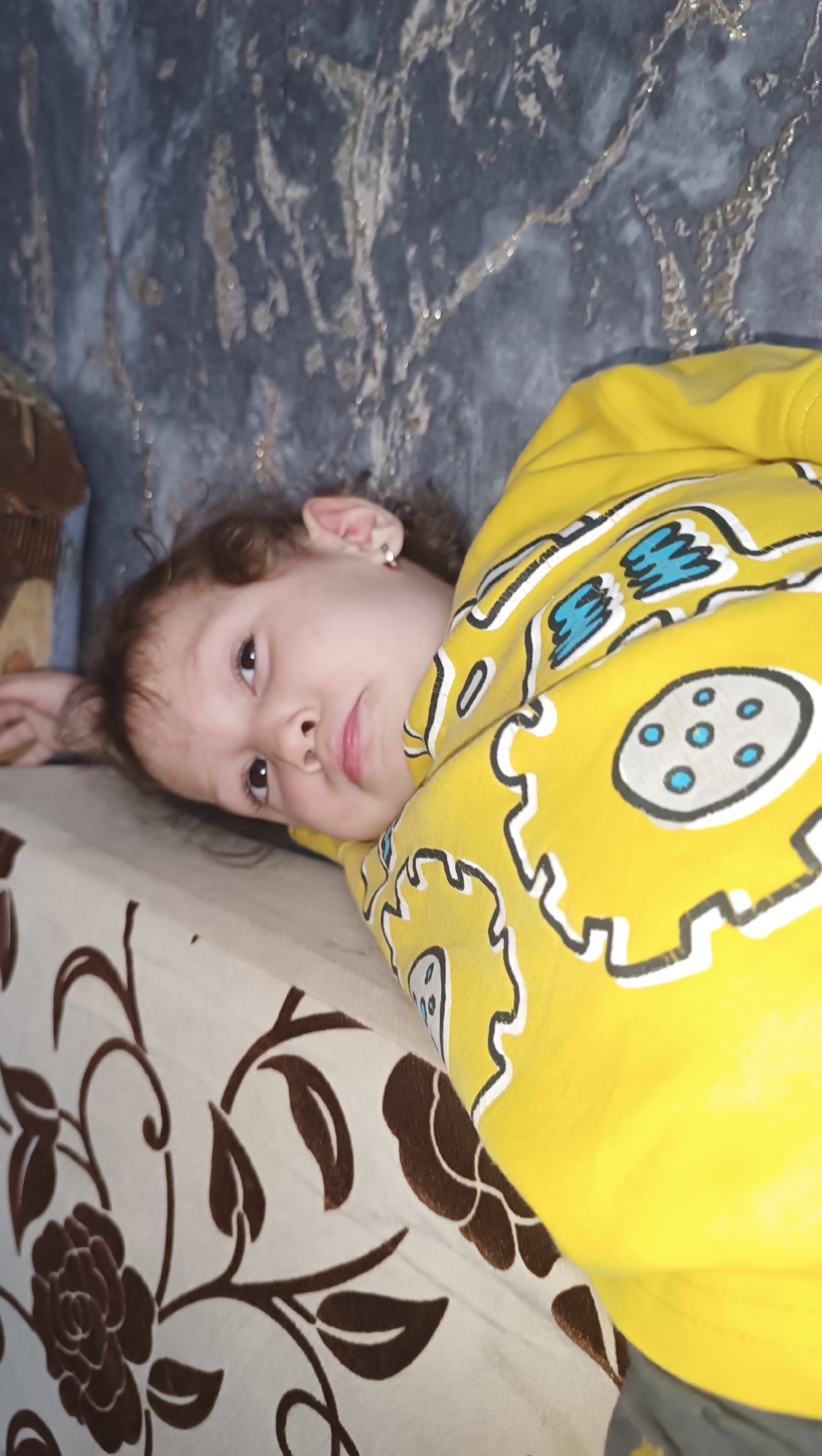 A young child, with curly brown hair and brown eyes, lies on their side on a beige surface with a brown floral pattern. The child is wearing a bright yellow sweatshirt with a graphic design featuring stylized blue and white circular shapes outlined in black. The child's right ear is visible, adorned with a small gold earring. In the background, a textured surface, possibly a wall, displays a gray and white marbled pattern. The scene appears to be indoors, likely in a domestic setting within the city of Karsha, Russia.