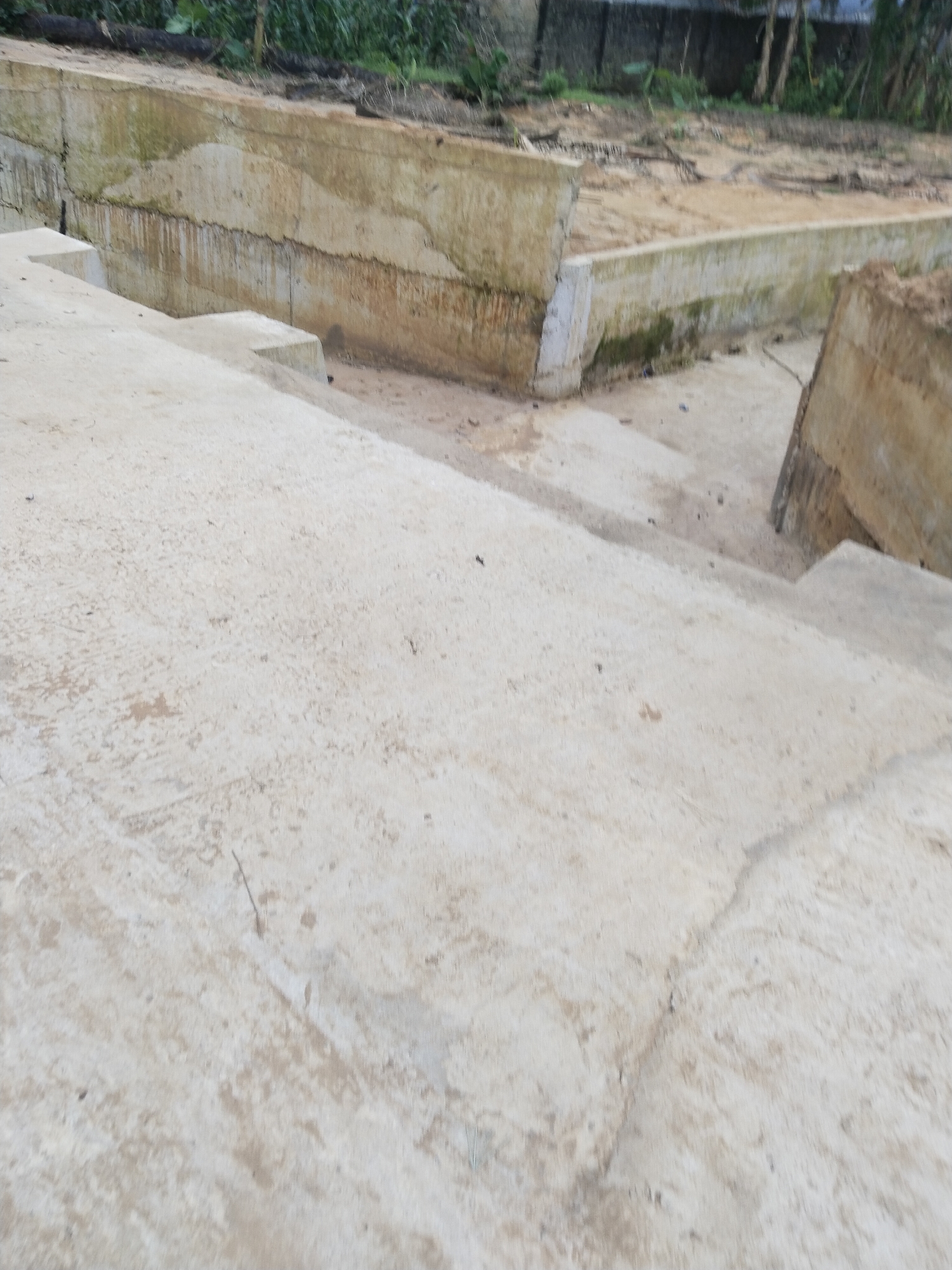 The image displays an outdoor scene dominated by concrete structures, suggesting a construction site or an area undergoing development. In the foreground, a large expanse of light-colored, rough concrete paving slopes gently, with several concrete steps descending on the left side. This concrete surface shows minor stains, dirt, and light cracking.
Towards the middle ground, a series of low concrete walls or retaining structures form boundaries. One prominent wall runs horizontally across the frame, exhibiting noticeable staining, streaks, and patches of what appears to be dirt or mold, indicating weathering and age. Behind this wall lies an excavated or disturbed area of light brown, loose earth or sand, appearing uneven. Another section of concrete wall, partially broken, is visible on the far right.
In the background, beyond the excavated ground, there is dense green vegetation and some blurred elements that could be distant structures or a fence. The lighting is diffuse and soft, consistent with an overcast day or early morning/late afternoon, as there are no distinct shadows. No individuals are present in the image, and no specific activity is taking place. The scene conveys a sense of ongoing or paused construction, possibly for a building foundation or a drainage system. No text is visible anywhere in the image. The setting aligns with an infrastructure development site in Uyo, Nigeria.