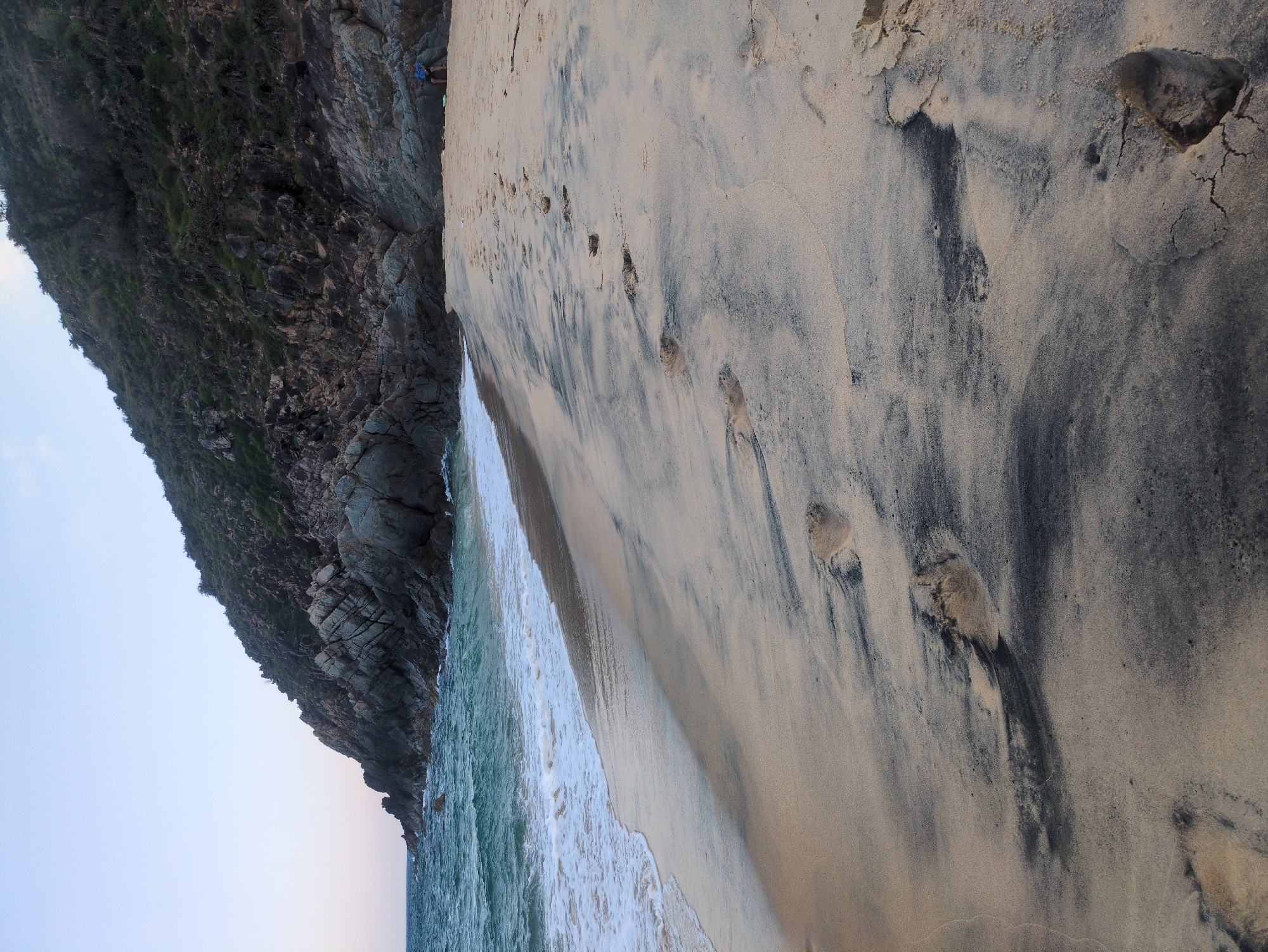 This image depicts a coastal scene in East Quy Nhon Ward, Vietnam. The upper frame shows a clear, pale blue sky. The ocean, characterized by turquoise-green water and gentle white-foamed waves, borders the left side of the scene. A steep, dark grey rocky cliff, partially covered with sparse dark green vegetation, dominates the right background and meets the ocean. The foreground and mid-ground are occupied by a sandy beach composed of light beige/tan sand distinctly mixed with dark grey/black sand, creating mottled patterns. Numerous barefoot human footprints are impressed into the sand, forming a trail from the mid-ground towards the foreground, with some showing cracked edges. On the far right, positioned at the base of the rocky cliff, a single individual is seated or crouching. This person wears a blue patterned shirt and dark shorts. A light-colored object rests on the sand immediately in front of the individual. No other people, structures, or significant activities are present.