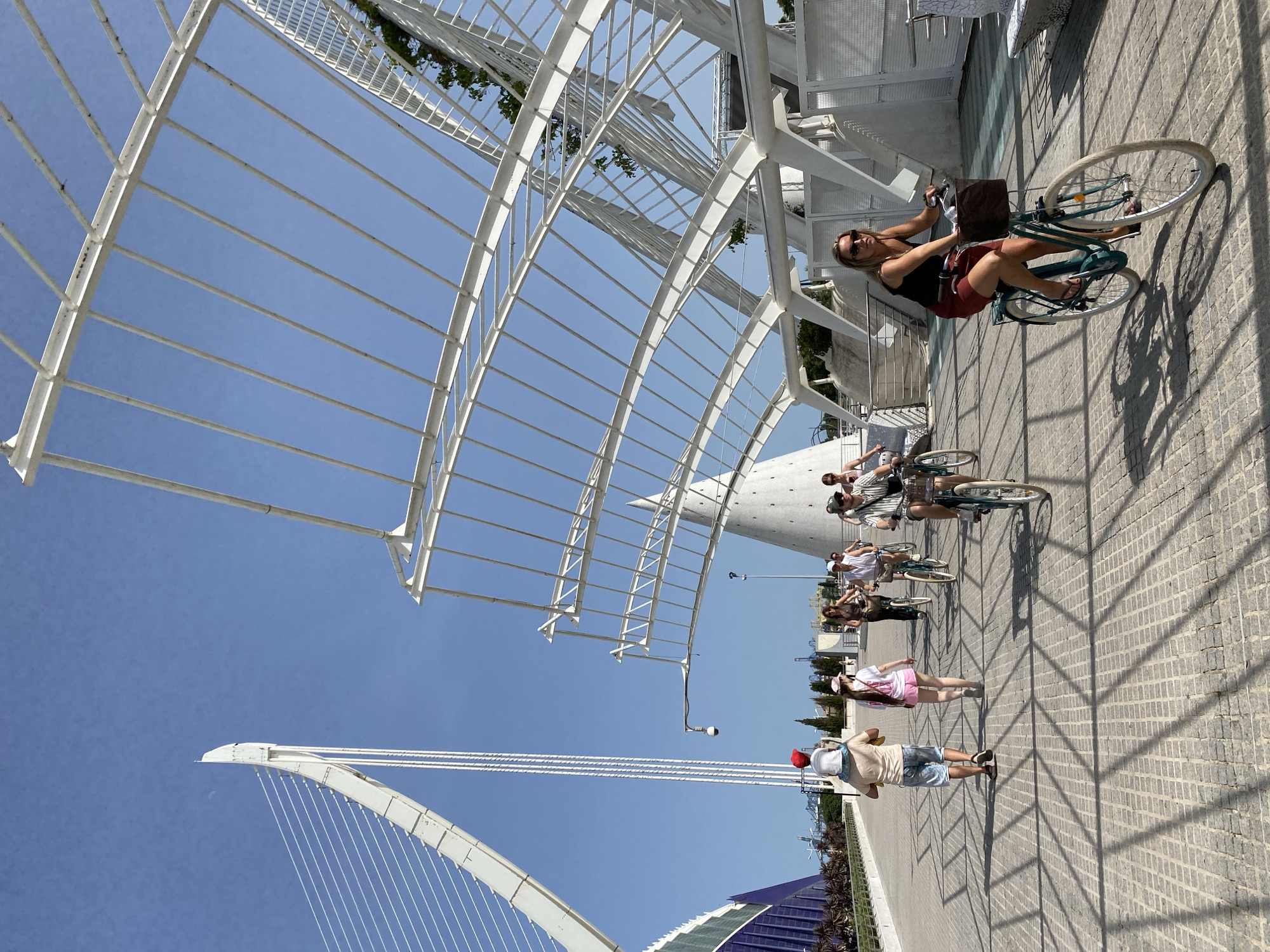 A group of people are walking and cycling on a paved walkway in a modern architectural area on a sunny day. In the foreground, a woman is riding a teal-colored bicycle with a basket in front, wearing sunglasses, a black tank top, and a red skirt. To her left, a man is walking with a child on his shoulders, wearing a red hat, a light-colored t-shirt, and blue shorts. Behind them, other individuals are walking and cycling. The background features a large, white, cantilevered structure with a blue building on the left and a conical structure. The walkway is paved with light-colored rectangular stones and exhibits a grid-like shadow pattern from the overhead structures. The sky is clear and blue. This scene is likely located in the City of Arts and Sciences in Valencia, Spain, known for its futuristic architecture.