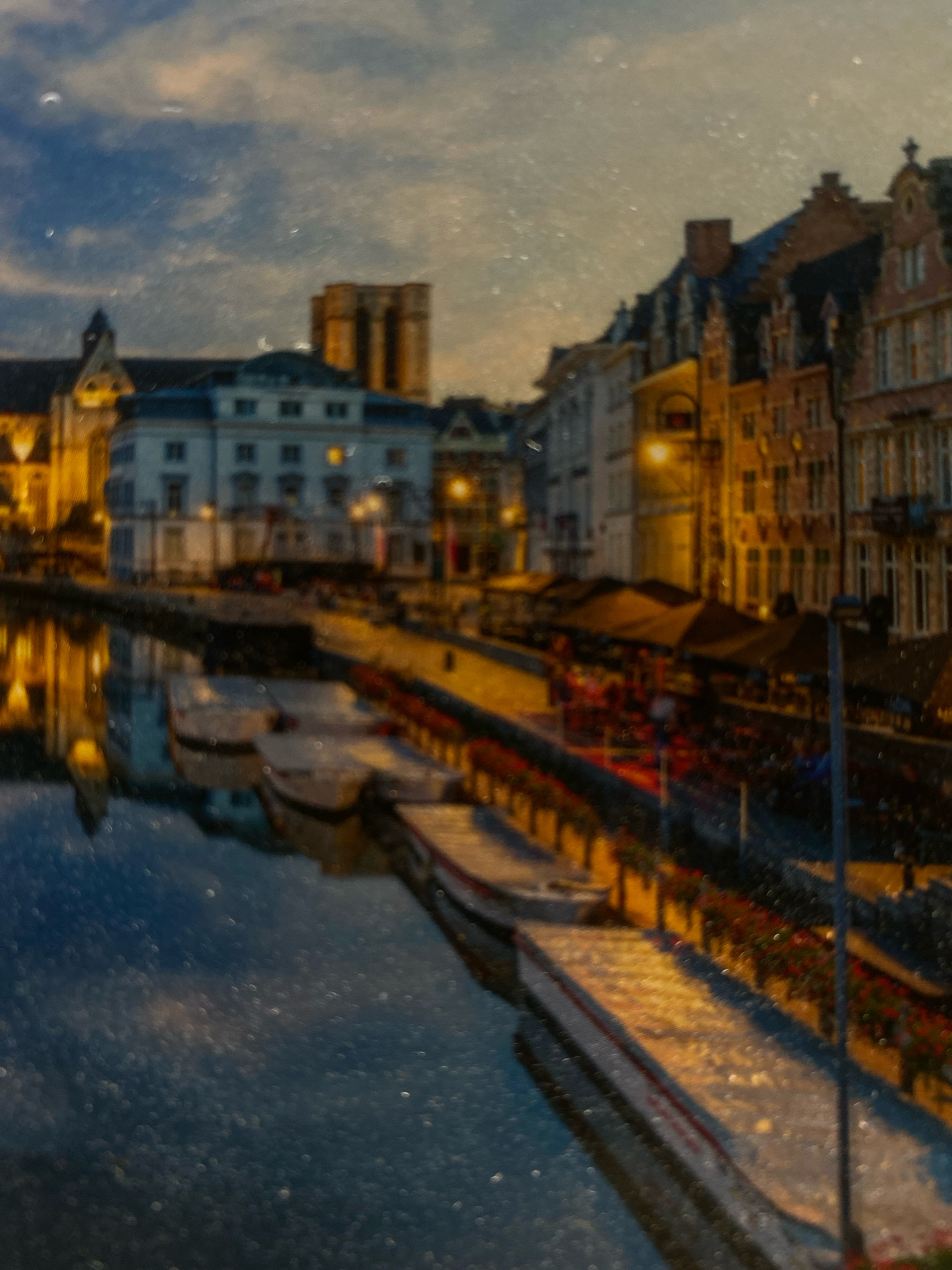 The image depicts a waterside promenade lined with buildings and outdoor seating areas. Several boats are moored along the water's edge. The scene appears to be illuminated by artificial lighting, suggesting it is evening or night. The architecture of the buildings suggests a European setting, with historical-style facades. There are no people readily visible. The water reflects the lights and buildings. The sky above is overcast. The location is not Zarechny, Russia.