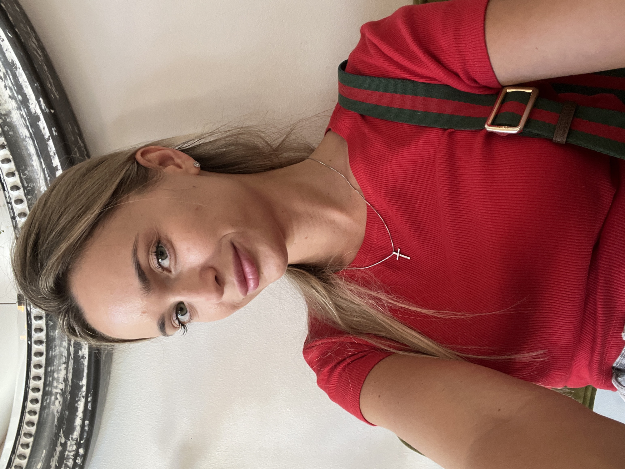 A young woman with blonde hair and green eyes is photographed from the chest up. She is wearing a red, ribbed t-shirt and a silver necklace with a cross pendant. A green and red striped strap with a gold buckle is visible over her right shoulder, suggesting she is wearing a bag. She is looking directly at the camera and smiling slightly. The background is a plain, off-white wall, with a decorative, ornate mirror partially visible in the upper left corner. The lighting suggests an indoor setting. The image does not contain any information about the specific location in Odesa, Ukraine.