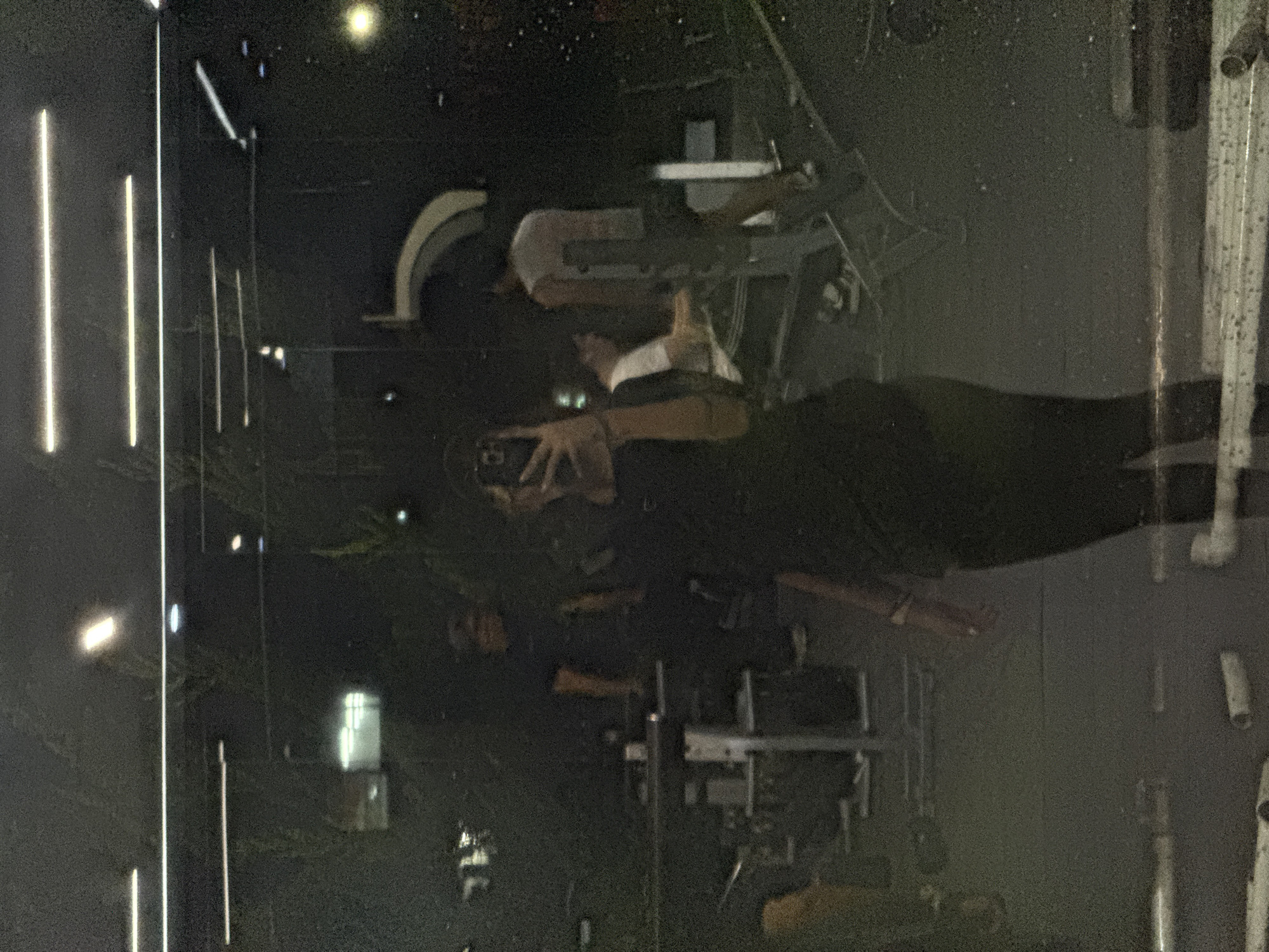 The image shows a person standing inside a building, reflected in glass. The individual is holding a phone, seemingly taking a picture of the reflection. The reflection shows a street at night with illuminated light posts and what appears to be building details. The reflection suggests the location is San Andrés Cholula, Mexico, based on architectural and urban characteristics observed. The scene is dark, with the reflection providing the primary light source.