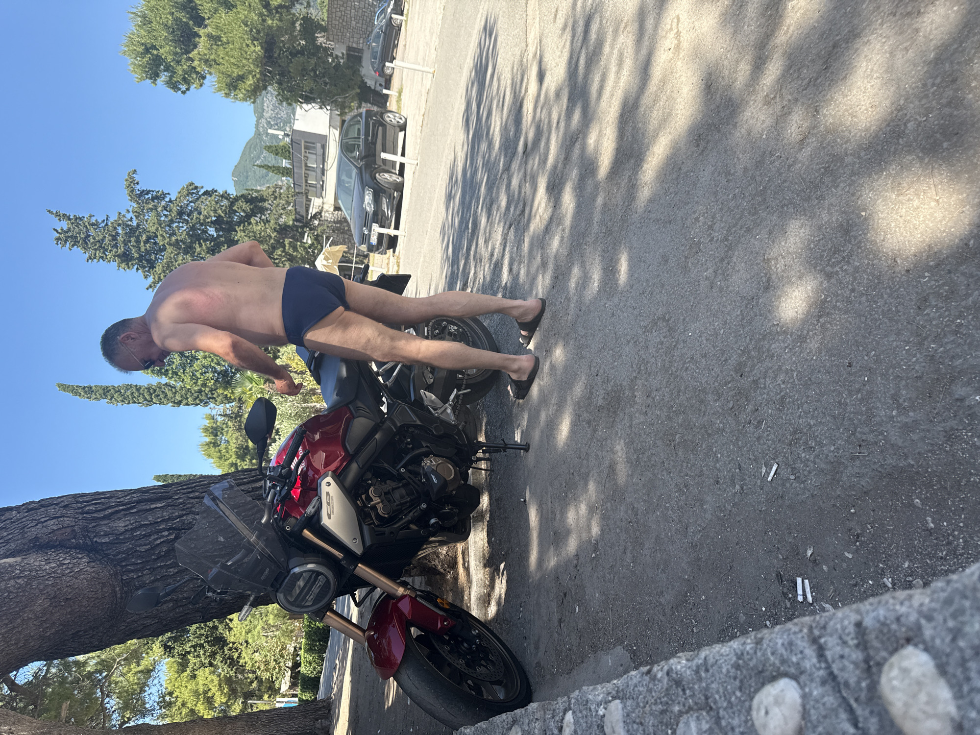 A man in dark blue swim trunks stands next to a red motorcycle parked under a large tree. The man is facing away from the camera, and appears to be adjusting his trunks. The motorcycle is a modern sport-bike with a dark tinted windshield. In the background, cars are parked along a road, and buildings with windows are visible. Beyond the buildings, mountains covered in trees rise under a clear blue sky. The ground is paved with asphalt, and there are some scattered cigarette butts. The scene is set on a sunny day in Budva, Montenegro, likely near a beach or recreational area given the man's attire and the proximity to parked cars which may belong to tourists. The overall impression is of a casual, summer day.