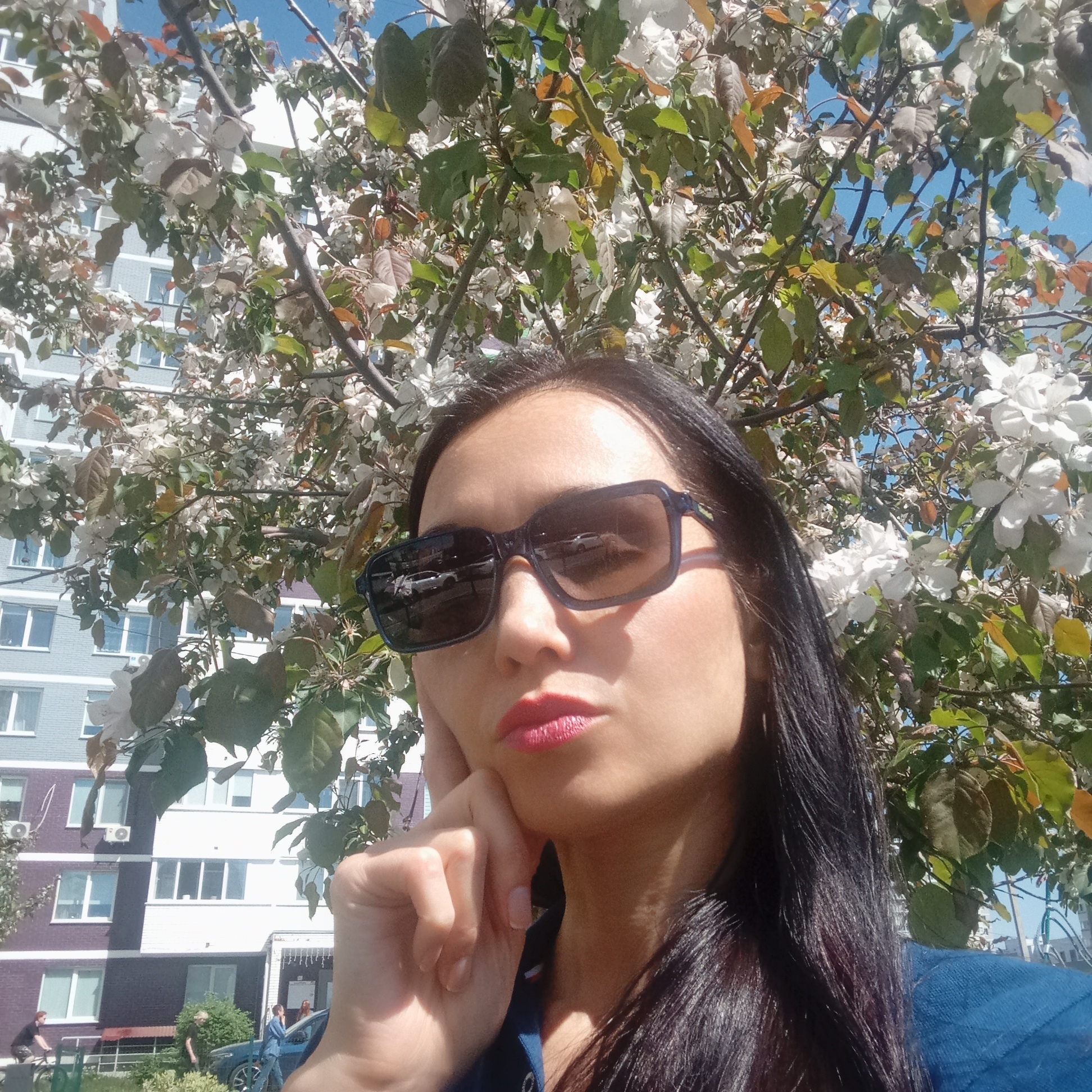A woman wearing sunglasses and red lipstick poses for a selfie in front of a flowering tree in Izhevsk, Russia. The background features a multi-story building with a facade painted in sections of white, light blue, and dark purple. In the lower left corner of the frame, glimpses of a bench, a parked car, and pedestrians are visible. The overall scene is taken in daylight with fair weather and likely in springtime given the blossoms. The subject is wearing a blue garment.