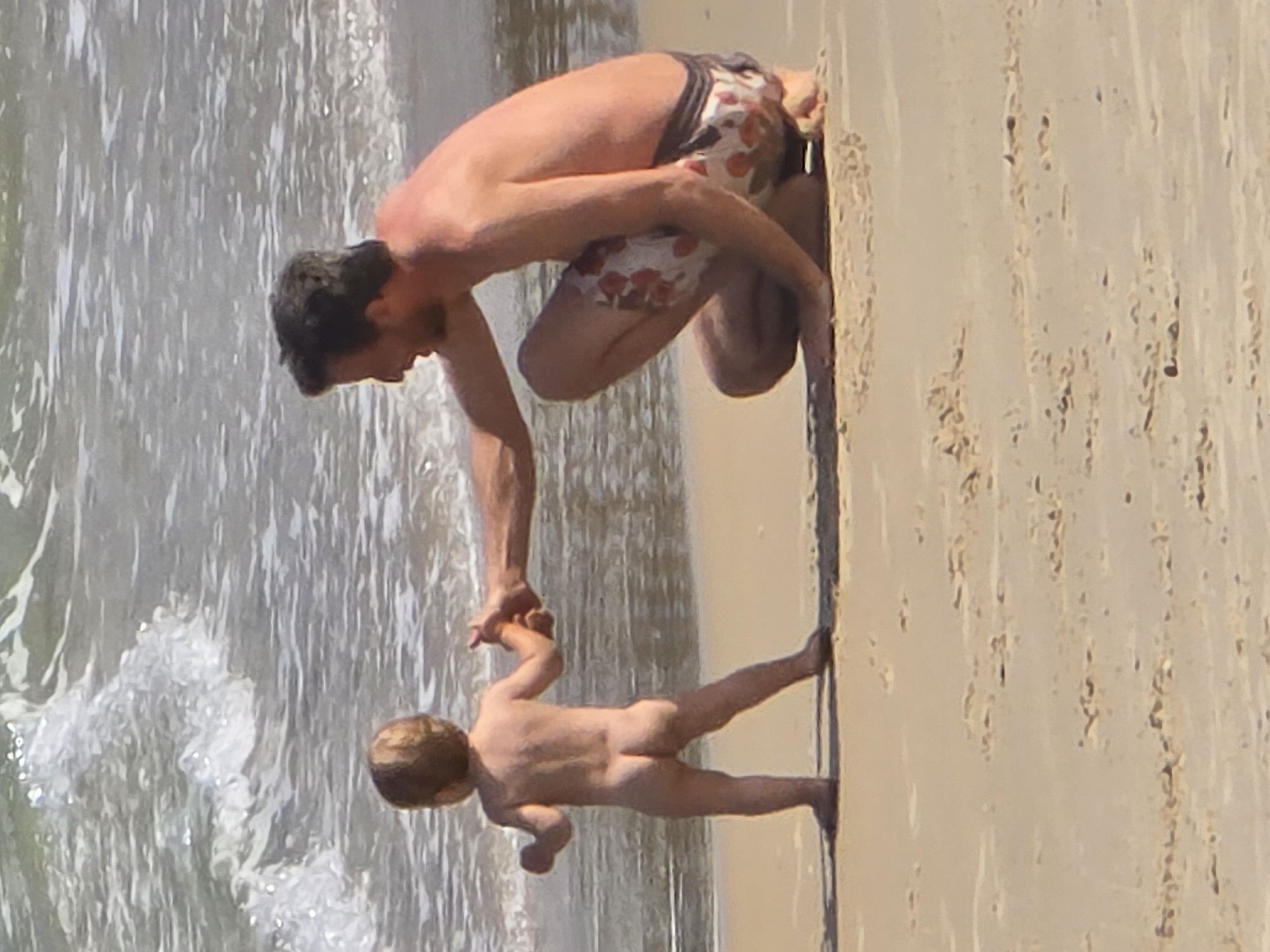 A man and a child are standing on a beach in Tel-Aviv, Israel. The man is wearing floral swimming trunks. The child is unclothed. The man is holding the child's hand, and they are both facing the water. The water is foamy and white. The sand is light brown.