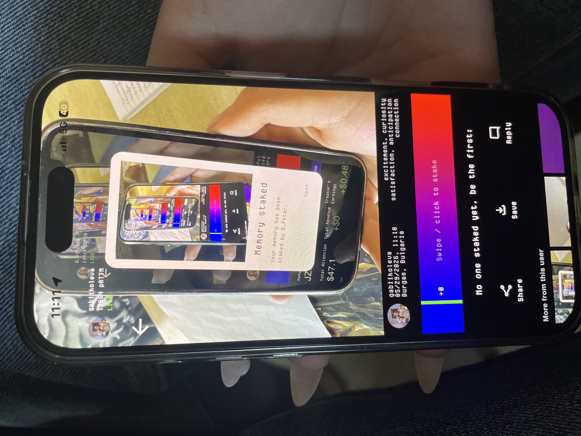 The image shows a person holding a smartphone, which displays a screen mirroring another instance of the same application on another phone. The application seems to be related to social media or content creation, with elements such as "Memory Staked," "Total Attention," and "Creator's Earnings" visible. The username "gabiikoleva" is displayed multiple times, alongside a timestamp "05/29/2026, 11:10" and the location "Burgas, Bulgaria." Additional textual elements like "excitement, curiosity, satisfaction, anticipation, connection" are present. The phone indicates a "5G" connection with a signal strength indicator. The person holding the phone is in a seated position on denim. The screen also shows the time "11:11."