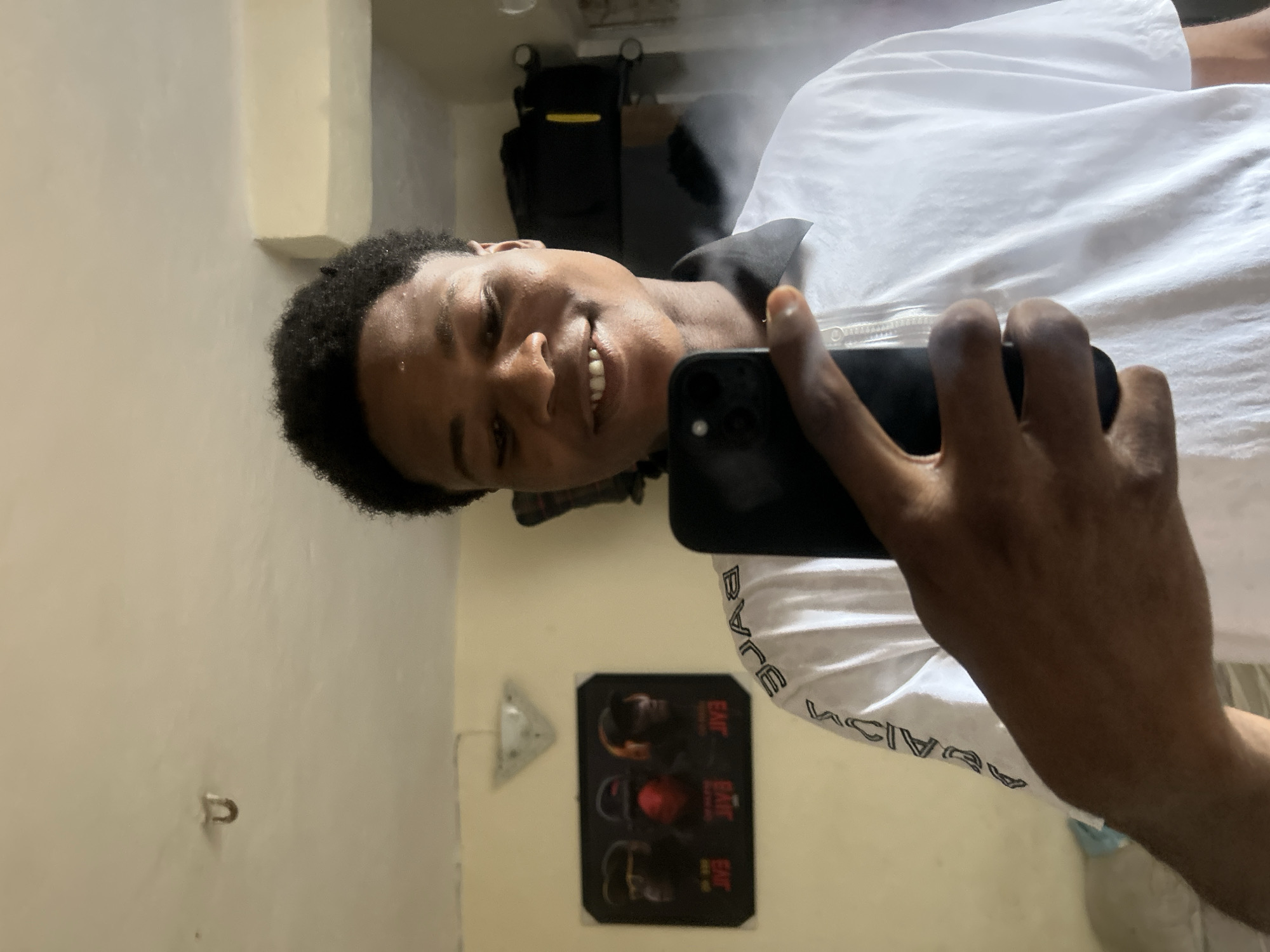 A young Black male, approximately in his late teens or early twenties, with short, dark, curly hair, is depicted taking a selfie with a dark-colored smartphone. He is smiling, revealing his teeth, and a slight sheen is visible on his forehead. He is wearing a white short-sleeved t-shirt with a black collar and a zippered front closure. The text "BAPE" is visibly mirrored on the left sleeve of his shirt. The setting is an indoor room in Ihiagwa, Nigeria, characterized by light-colored, possibly cream or beige, walls and an off-white ceiling. A U-shaped metal hook is affixed to the ceiling. On the wall to the viewer's left, a framed poster features three stylized figures, each with mirrored "BAPE" text below them, positioned beneath a triangular-shaped light fixture. To the viewer's right and behind the subject, a dark bag or piece of luggage with a yellow accent is visible. Below the bag, a localized white haze or mist is present.