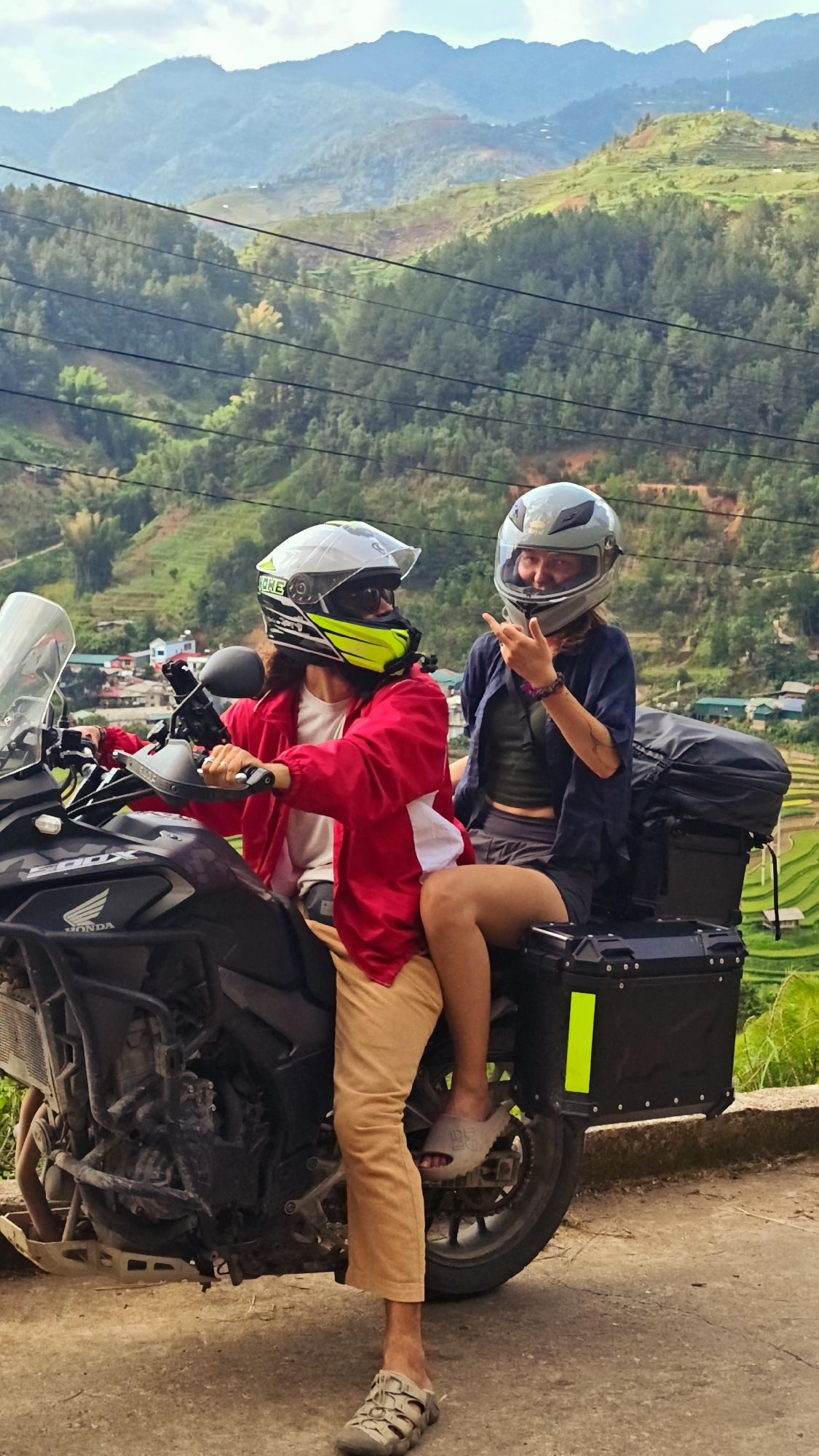 This image features two individuals on a motorcycle, stopped on a paved surface, against a breathtaking mountainous backdrop. The scene suggests a scenic stop during a journey in a rural, elevated region, consistent with the location context of Lao Chai, Vietnam, known for its terraced landscapes.
The driver, positioned on the left, wears a white full-face helmet with vibrant yellow accents and dark sunglasses, obscuring their face. They are dressed in a bright red jacket over a lighter shirt and light brown pants, with a grey sandal on their visible foot. The motorcycle is a black Honda, with "HONDA" clearly visible on its side and "500X" faintly discernible.
The passenger sits behind the driver on the right, wearing a grey full-face helmet. She has a bright smile and is making a two-finger gesture (possibly a peace sign) with her right hand, indicating a cheerful and relaxed mood. She wears a dark blue open shirt over a dark top and shorts, with grey slippers on her feet. Behind her, a black hard-shell luggage box with a bright yellow reflective strip is attached to the motorcycle, along with another soft black bag.
The background showcases lush green mountains with distinct rice terraces carved into the slopes, interspersed with dense tree cover. Small clusters of buildings are visible in the distance. Multiple power lines run horizontally across the mid-ground. The sky is bright with some clouds, suggesting daytime conditions. Visible text includes "HONDA" on the motorcycle and "CORE" on the driver's helmet. Some indistinct text appears on the passenger's slipper.