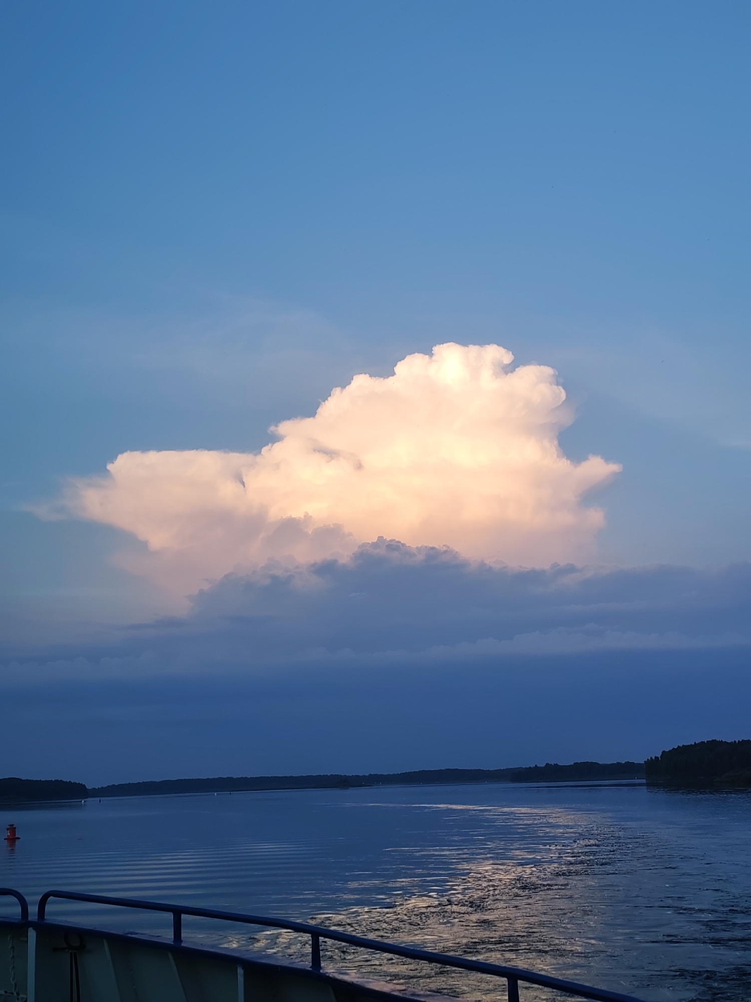 The image captures a serene scene of a body of water, likely a river or a bay, with a distant tree-lined shore. A large, luminous cumulus cloud dominates the upper half of the frame, tinged with warm orange and pink hues, suggesting it is either sunrise or sunset. Below the prominent cloud, darker, lower-lying clouds create a layered effect against the deep blue sky.
The water's surface is calm, with gentle ripples reflecting the sky's colors and the fading light. A faint wake trails behind a vessel from which the photo appears to be taken, indicated by the presence of a railing in the foreground. A solitary red buoy is visible on the left side of the water, marking a channel or hazard.
The overall mood is peaceful and tranquil, evoking a sense of calm and natural beauty. The time of day is clearly either early morning or late evening, with the low-angled light illuminating the clouds. The location is identified as Ul'yanovo, Russia, which aligns with the natural landscape depicted. No text is visible in the image.