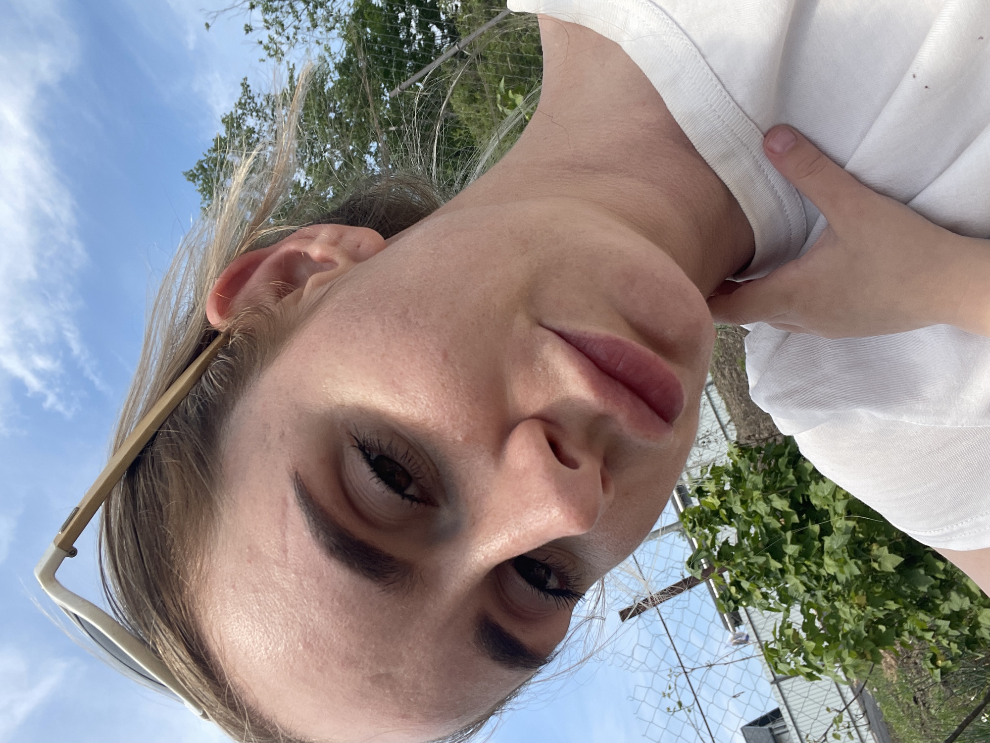 A close-up, angled selfie of a fair-skinned woman with light brown hair. She is wearing a white t-shirt and sunglasses, with the frame visible on the left side of the image. Her face is the primary focus. The background includes a blue sky with clouds, green foliage, a chain-link fence, and glimpses of a building structure. There's no information to indicate the image was taken in Yasnoe, Russia.