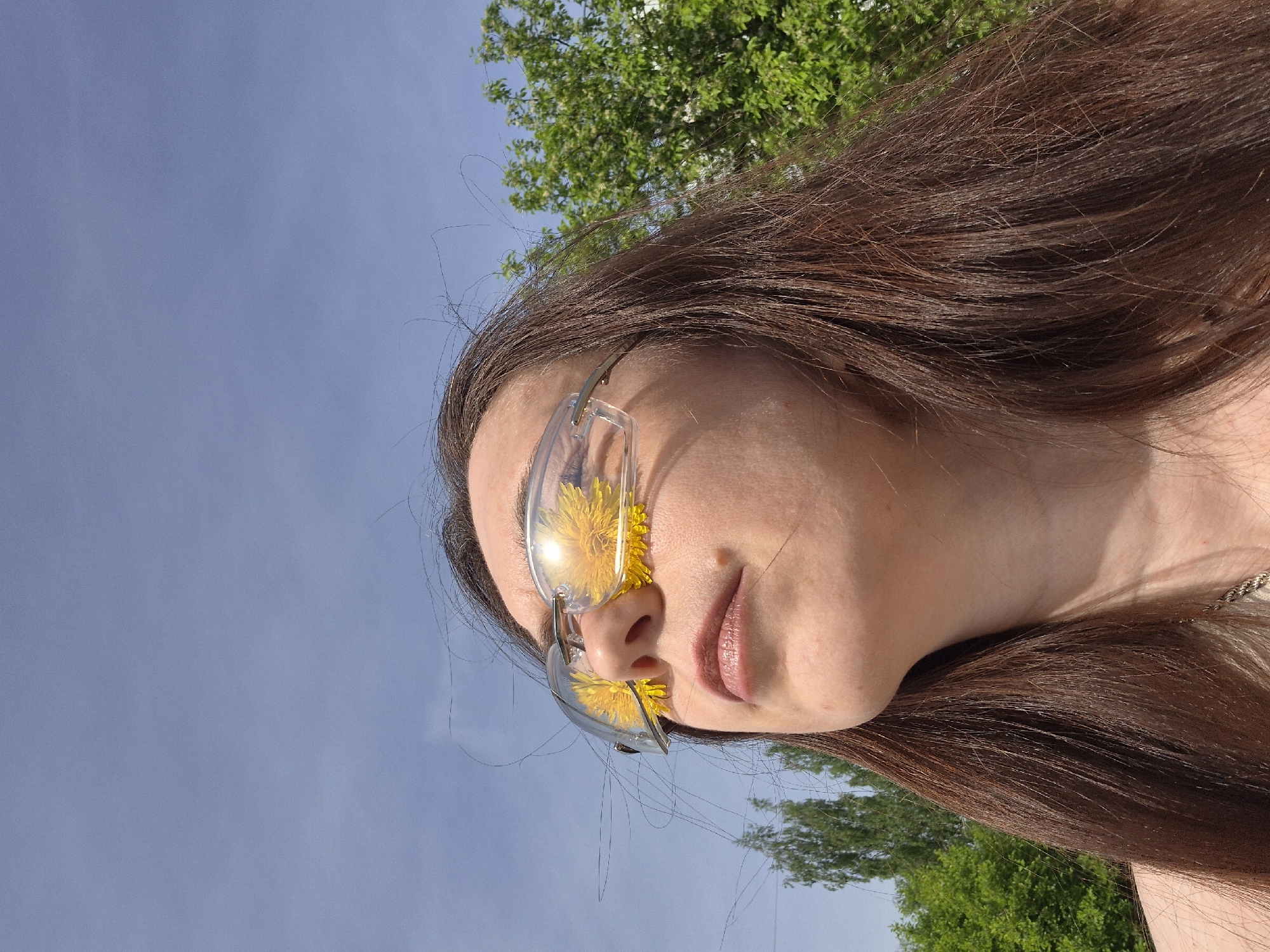 A low-angle, daytime outdoor shot shows a female with long, brown hair wearing eyeglasses with yellow flowers placed inside the lenses. The woman's face is tilted slightly, and she has light skin. A hint of a necklace is visible on her neck. The background consists of a blue sky and green tree foliage. Location is in the city of Unknown, Unknown.