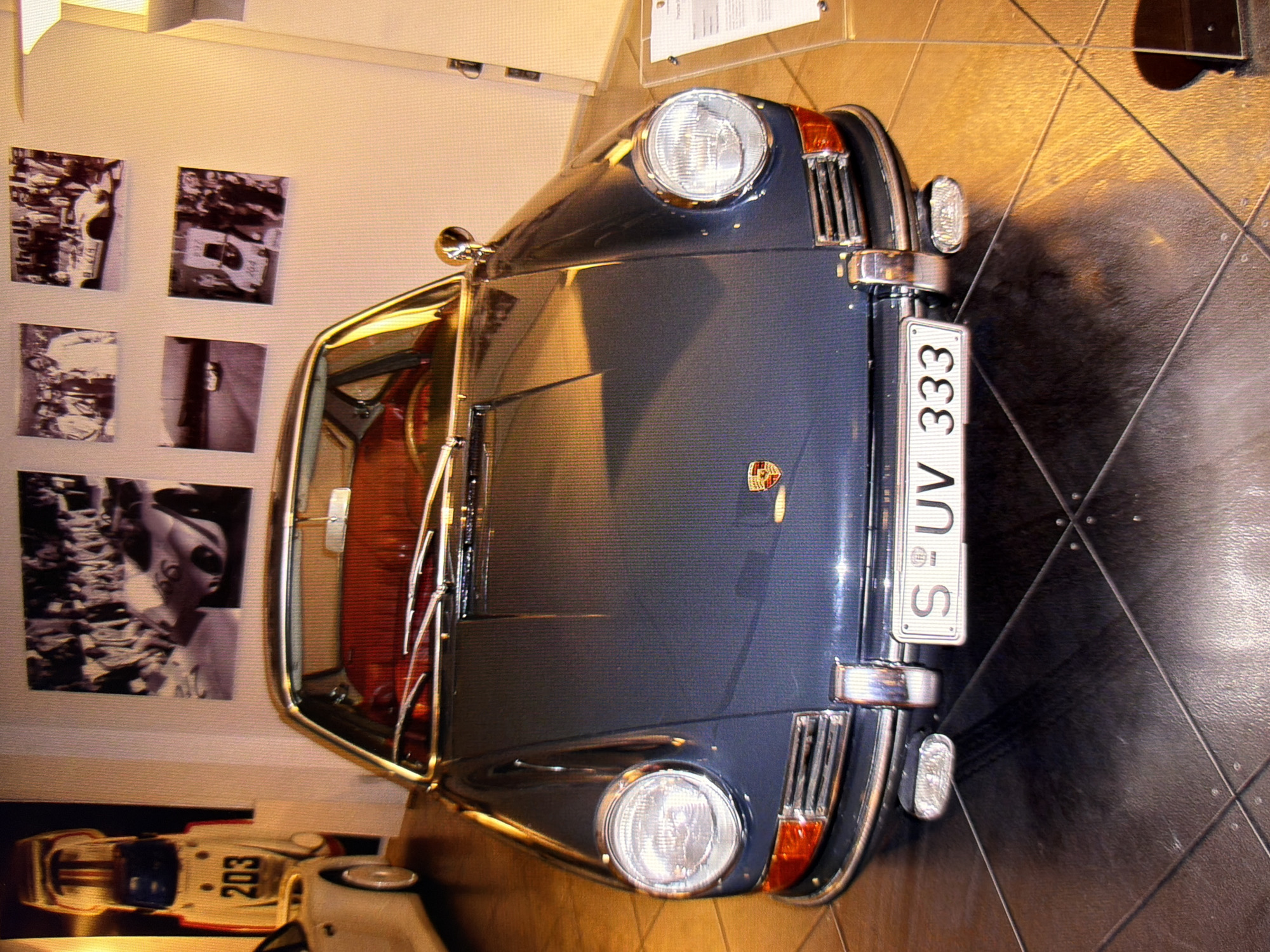 The image depicts the front view of a dark blue vintage Porsche 911 parked indoors, likely in a museum or showroom. The car is the central focus, with its headlights, grille, and license plate clearly visible. The license plate reads "S-UV 333". Behind the car, on the wall, are several framed black and white photographs, some featuring vintage racing cars and crowds. The floor appears to be tiled. To the right, a display stand with a white document is partially visible. The lighting suggests an indoor environment with artificial illumination. The scene is devoid of people. There is no indication of the specific location within the city of Kyiv, Ukraine.
