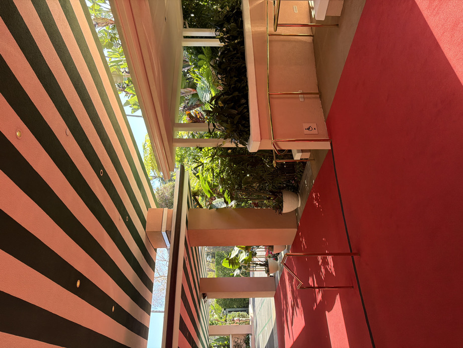 A low-angle perspective shows the exterior of a building with a striped pink and black wall. A pink awning extends from the building, framing a view of lush greenery and foliage. A red carpet leads up to a set of steps with gold railings, and the wall beside the steps features a sign indicating accessible facilities. The scene suggests a welcoming and accessible entrance to a building surrounded by nature.