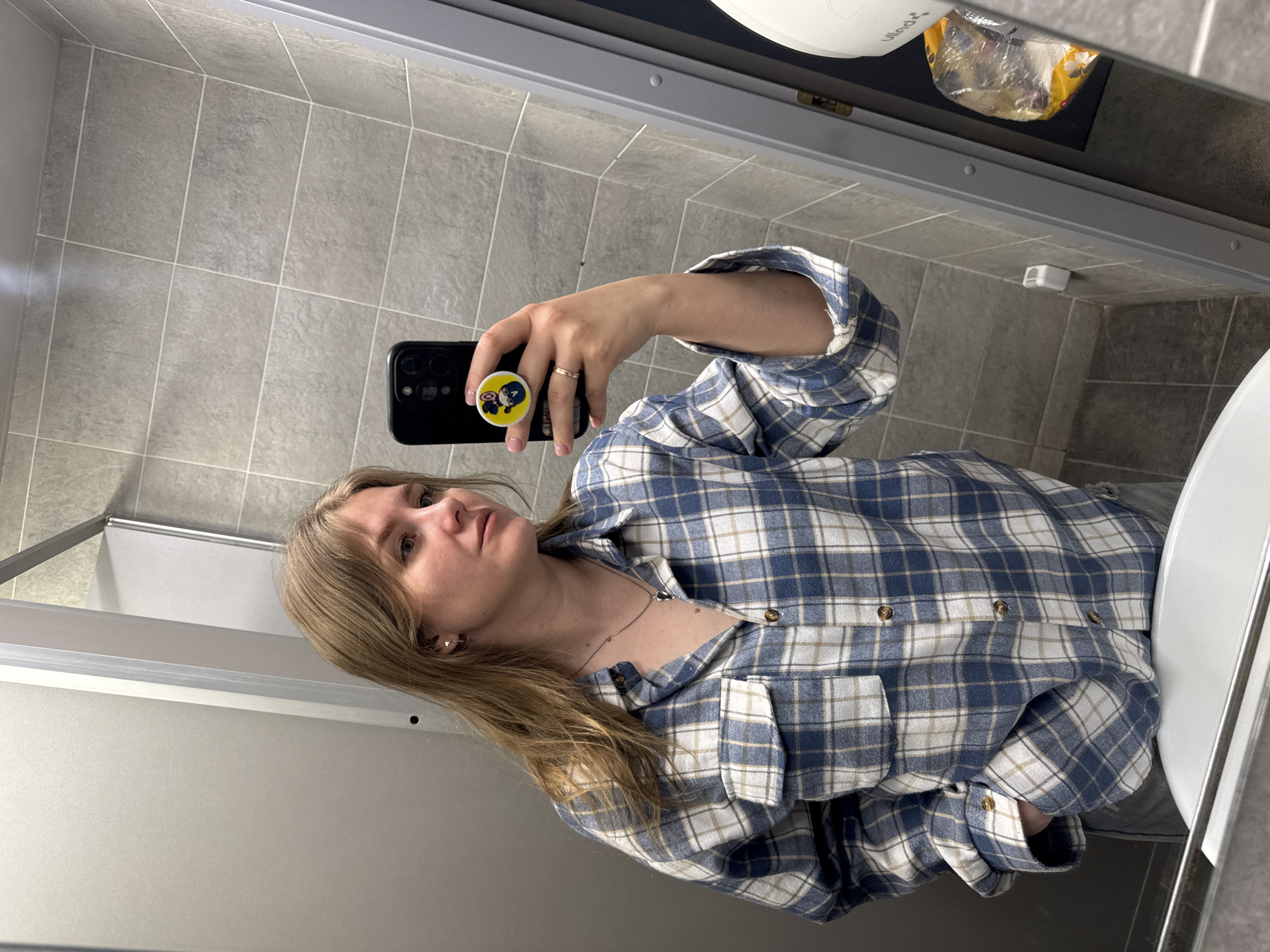 A mirror selfie taken in a restroom, featuring a woman with shoulder-length blonde hair wearing a blue and white plaid flannel shirt. She is holding a black smartphone with a yellow and blue Captain America pop socket. The restroom has gray tiled walls and floors, and a white sink and toilet are visible. A package of sanitary napkins is seen above the mirror. The location in the city of Perm, Russia, is not evident from the provided image.