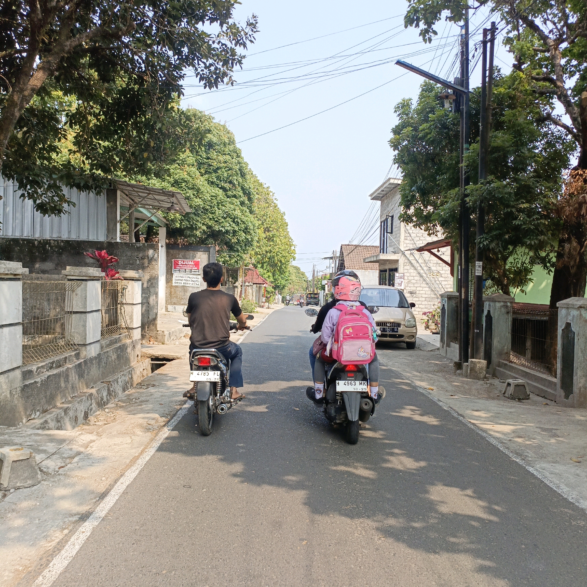 This is a rear-view, eye-level shot of two motorcycles traveling down a paved road in Salatiga, Indonesia. The motorcycle in the foreground carries a driver and a passenger, the latter wearing a pink helmet and a pink backpack adorned with cartoon characters. The motorcycle behind it has a single male driver wearing a dark t-shirt. Both motorcycles have license plates visible. The road is flanked by a sidewalk and a concrete barrier on the left, with houses and trees lining the right side of the road. Overhead, power lines are strung between utility poles. The scene suggests a typical daytime street view in a residential area.