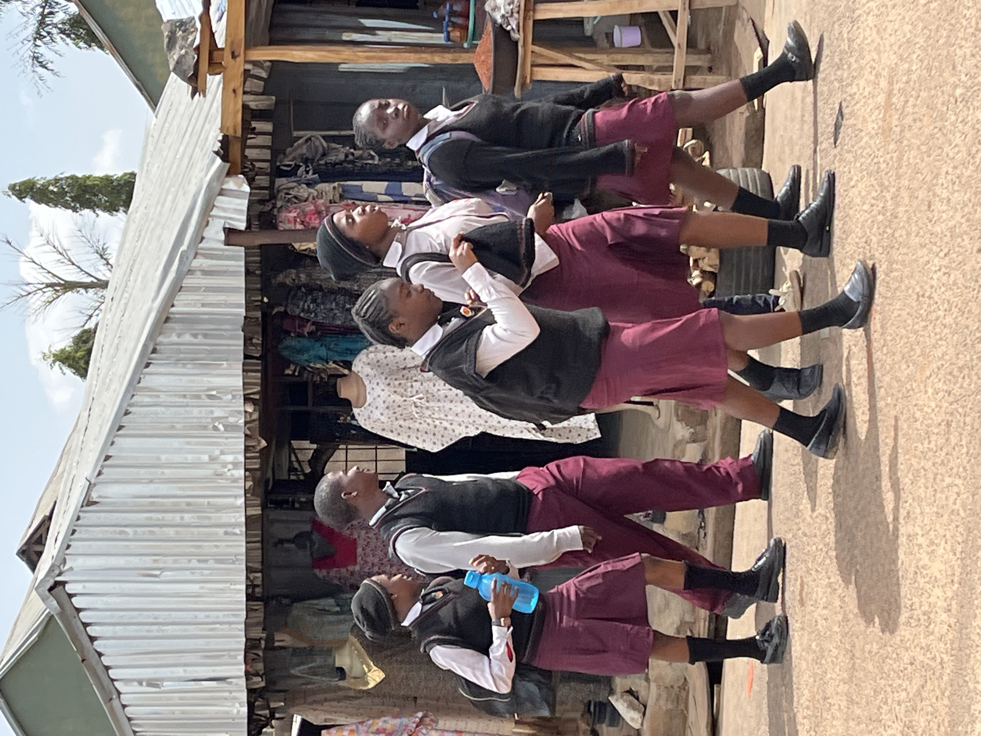 Five students, wearing school uniforms with maroon skirts or trousers, white shirts, and dark vests, stand in a line outside a building with a corrugated metal roof in Jos, Nigeria. The building appears to be a shop with clothing displayed. The students all face in the same direction, with the last student in line holding a blue water bottle.
