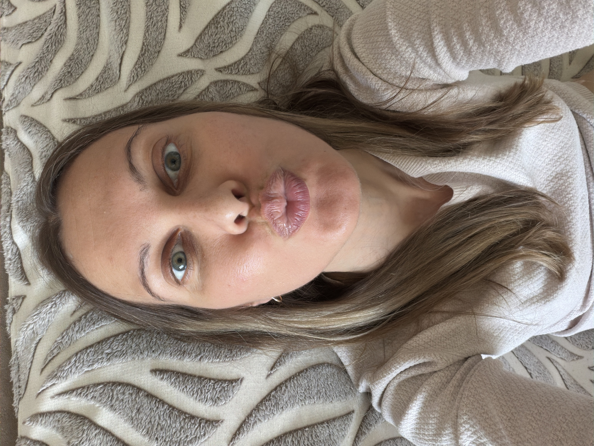 A woman with light brown hair and green eyes is taking a selfie. She is pouting her lips and looking at the camera. She is wearing a light pink sweater. She is lying down on a patterned surface that appears to be a pillow or blanket. The background is blurred. The setting appears to be indoors. The image was taken in Izhevsk, Russia.