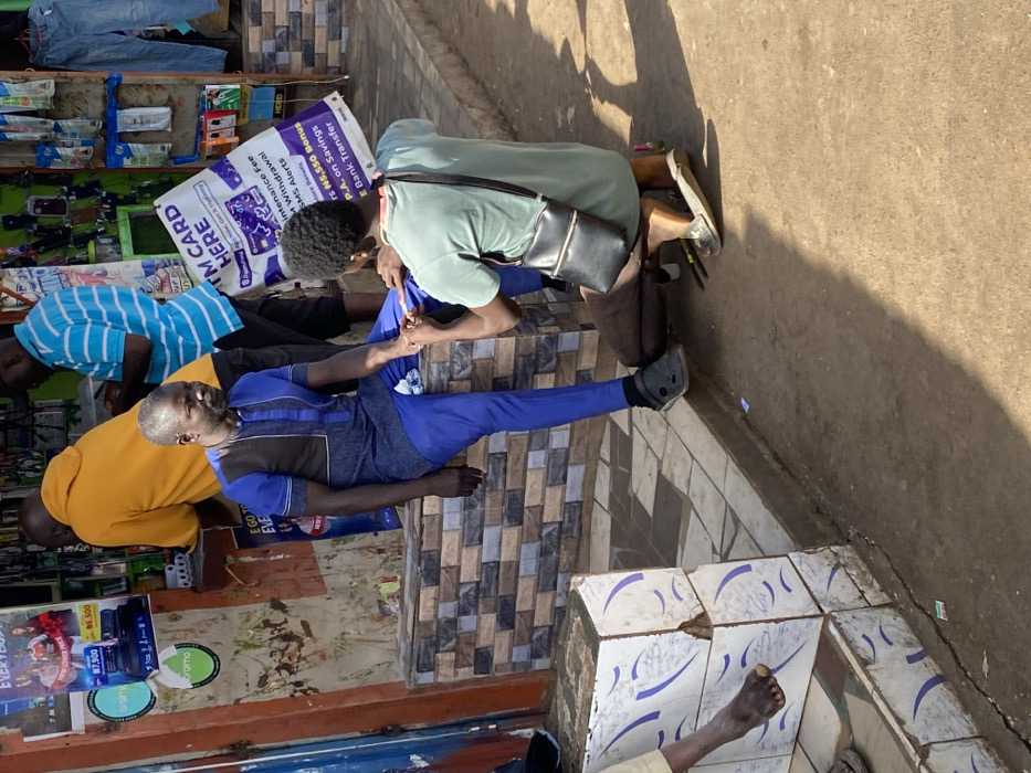 This image captures a street-level commercial area in Jos, Nigeria, under bright daylight. In the midground, a man with a grey beard, dressed in a blue and black patterned tunic and blue trousers, sits on a low, tiled bench. He wears dark slip-on shoes and looks towards the left. Kneeling before him on the bare earth ground is a younger individual, wearing a green t-shirt, brown trousers, and a black shoulder bag, attending to the seated man's right hand, indicative of a manicure or similar grooming service.
Behind the seated man, two other men are partially visible near the entrance of a shop. One wears a yellow hoodie, and the other a blue and white striped shirt. The shop's exterior wall is adorned with multiple advertisements, including green "oram" stickers and a prominent purple and white banner advertising "ATM CARD HERE" services, listing options like Paga, Palmpay, Opay, MTN Card, and detailing transactions such as withdrawals, SMS alerts, and bank transfers. Displays of what appear to be mobile phone accessories and electronics, marked with prices like ₦7,000 and ₦6,500, are visible on the shop wall. In the lower left foreground, a tiled platform shows a bare human foot resting on it. The immediate ground surface in front of the bench is bare earth.