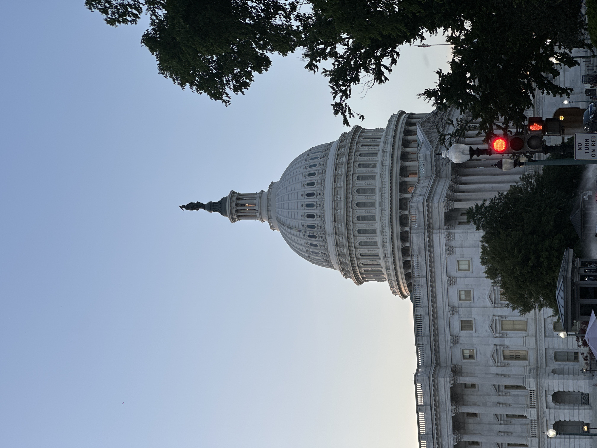 The image shows the United States Capitol Building in Washington, D.C. during daylight. The building's dome is prominently featured, and trees frame the upper portion of the image. A traffic light with a red light illuminated is visible to the right of the building. A "NO TURN ON RED" sign can be seen next to the traffic light. The sky is light blue.
