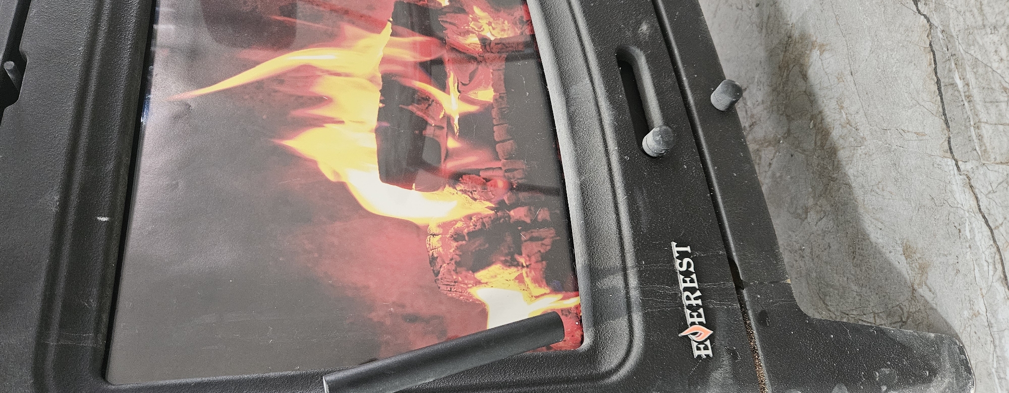 A close-up, eye-level shot shows the front of a black, electric fireplace, positioned on a light-colored, marbled floor. The fireplace features a glass door displaying a realistic image of burning logs and vibrant orange and yellow flames. A black handle is visible to the left of the glass door, and the brand "EVEREST" is embossed in silver with a small orange flame logo to the left of it, centered on the lower part of the fireplace. Two small, dark, circular knobs are present below the glass door. The overall scene is indoors, in what appears to be a residential setting, in the city of Semey, Kazakhstan.