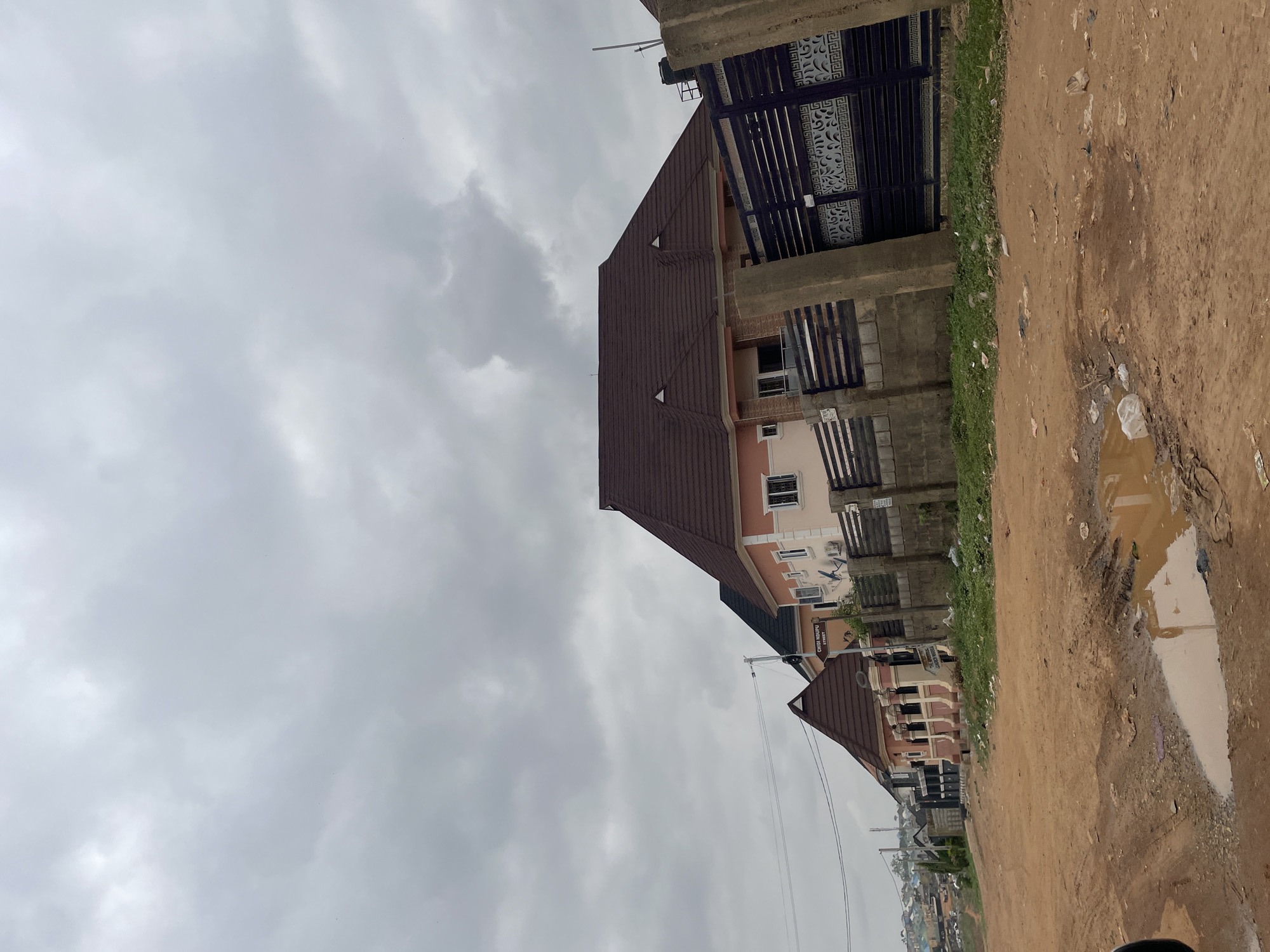 The image depicts a street in Abuja, Nigeria, under a cloudy sky. The street is unpaved with a puddle visible in the foreground. Several two-story buildings line the street, constructed of concrete blocks and painted in shades of beige and peach. The buildings have brown shingled roofs, and some feature ornate metal gates and balconies. Power lines are strung between the buildings. A street sign reading "CHIDI MOHANUI STREET" can be seen. The surrounding environment appears residential.