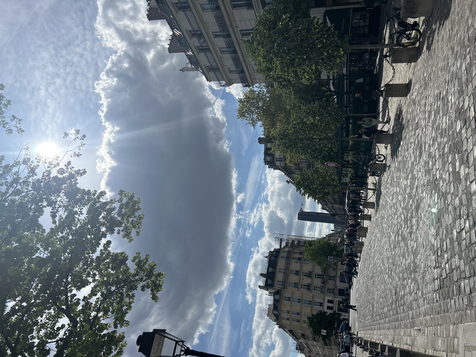 A street scene in Paris, France, featuring cobblestone pavement on the right and buildings on both sides. The sky above is filled with clouds, some dark and some bright, with the sun visible through the trees on the left. Along the street are trees, benches with people sitting on them, and bicycles parked along the edge. The Eiffel Tower can be seen in the distance between the buildings.