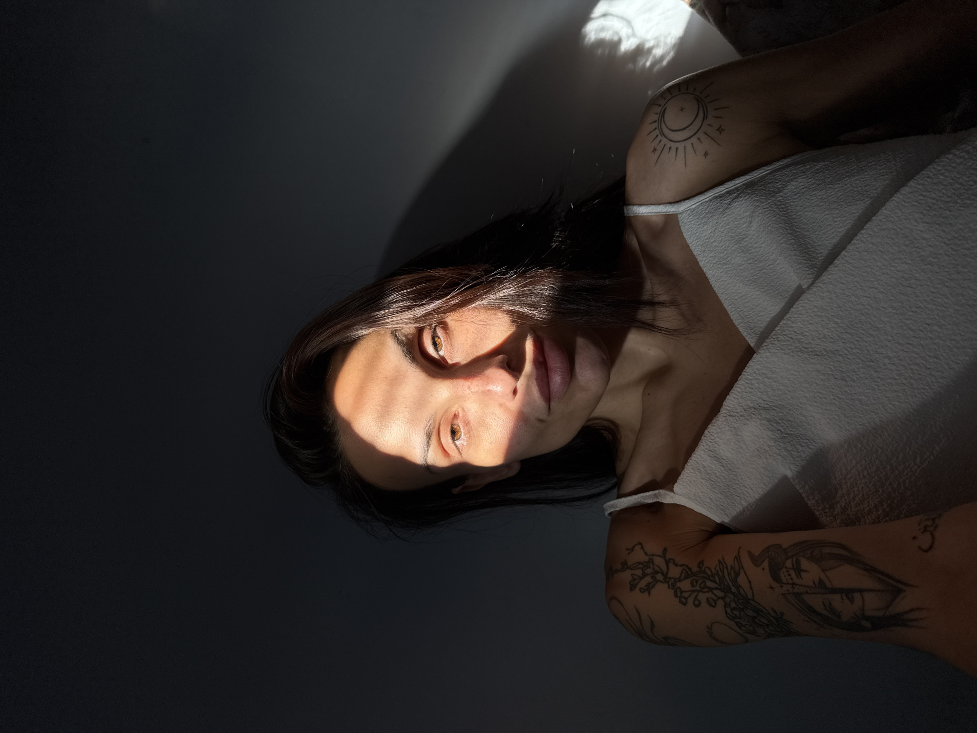 A portrait shows a person with long, dark hair and light skin. Light falls across the face from the upper left, illuminating part of the forehead, an eye, the nose, and upper lip. The person is wearing a white top. Visible tattoos adorn the right shoulder and arm. The background is a dark gray color. The location is not discernable, nor is there any visual evidence to indicate the location in Pleven, Bulgaria.
