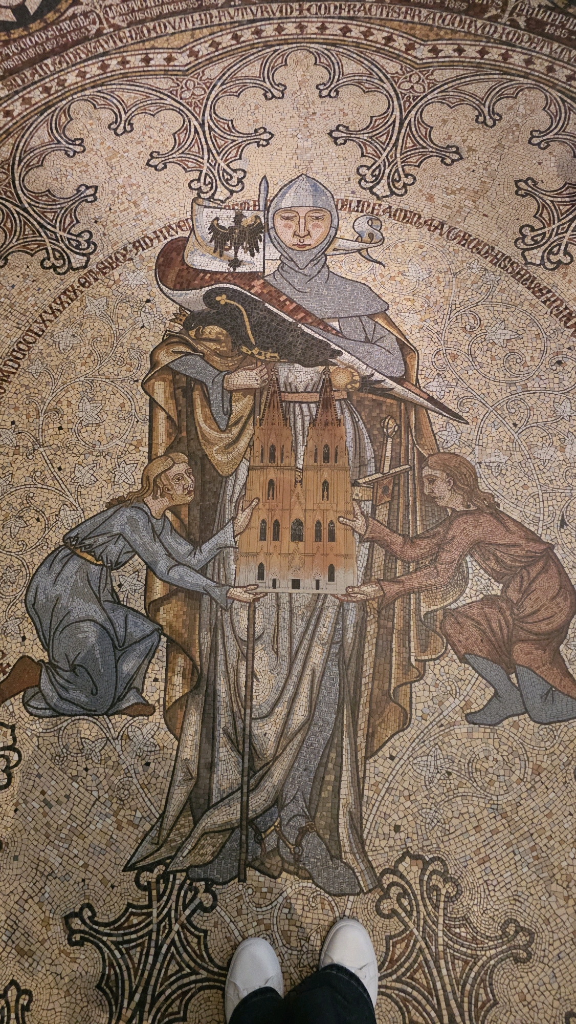 The image shows a mosaic floor depicting a central figure in a grey hooded robe, holding a model of a Gothic cathedral. The figure has a stern expression and wears a silver helmet. To the left, a kneeling figure in a blue robe holds up one side of the cathedral model with both hands. To the right, another figure in a brown robe kneels, reaching out to support the other side of the model with their hands. An eagle with spread wings is depicted on a shield behind the central figure's left shoulder, and a dark bird with a long beak is perched on the central figure's arm. The mosaic is intricate, with a textured background of small tiles. The floor appears to be part of an architectural space, possibly a historical building, given the style of the mosaic. The location is the floor of the Cologne Cathedral in Cologne, Germany.