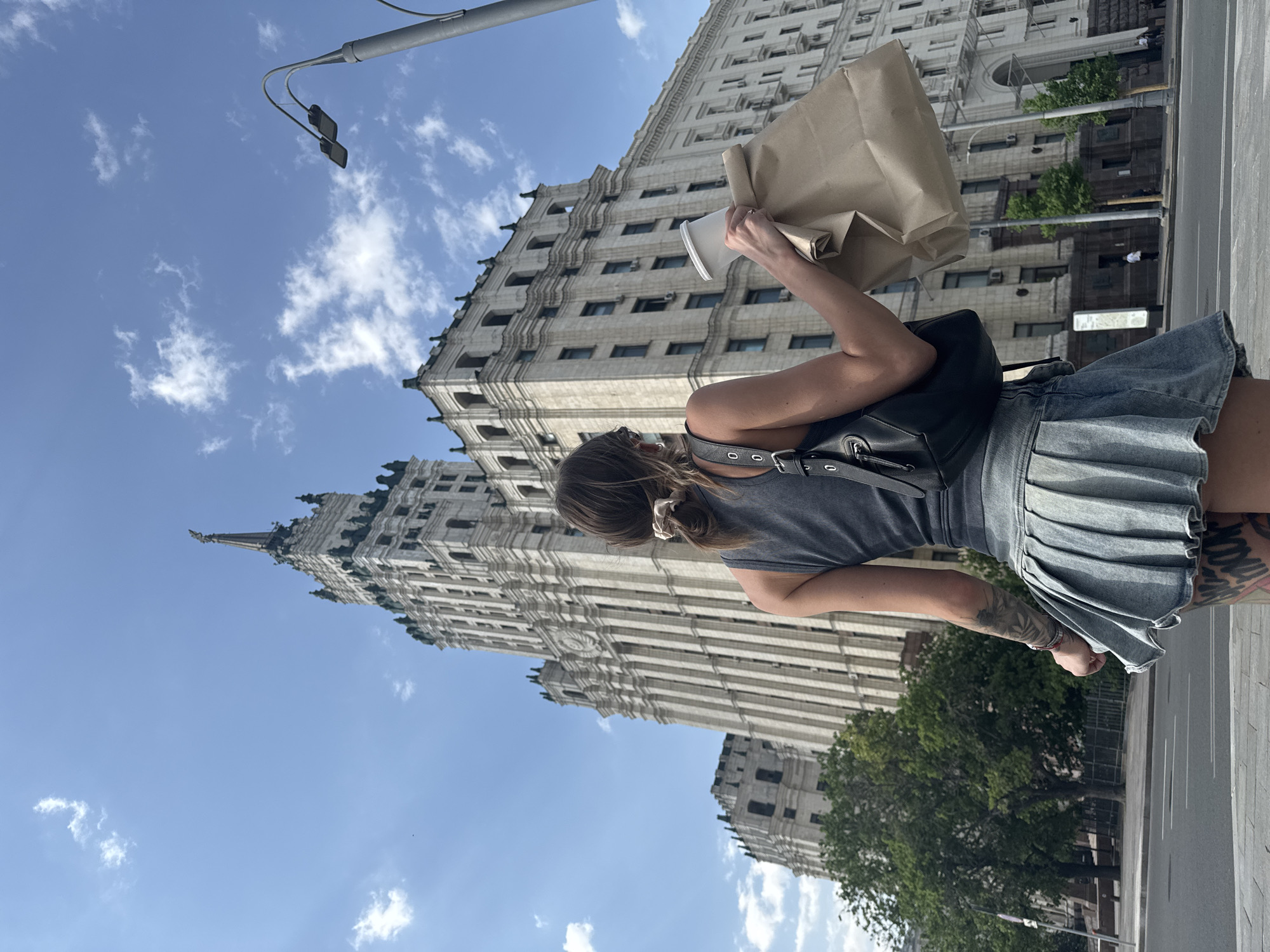 A medium-shot photograph displays a woman standing in Moscow, Russia, with the Kotelnicheskaya Embankment Building prominently in the background. The woman is holding a paper bag and a disposable cup in her hands. She is wearing a grey tank top, a grey denim skirt, and a black backpack. The woman has tattoos on her left arm. The building in the background is a tall, Soviet-era skyscraper with a distinctive spire. The sky is blue with scattered white clouds. A lamppost is visible in the upper left corner of the frame.