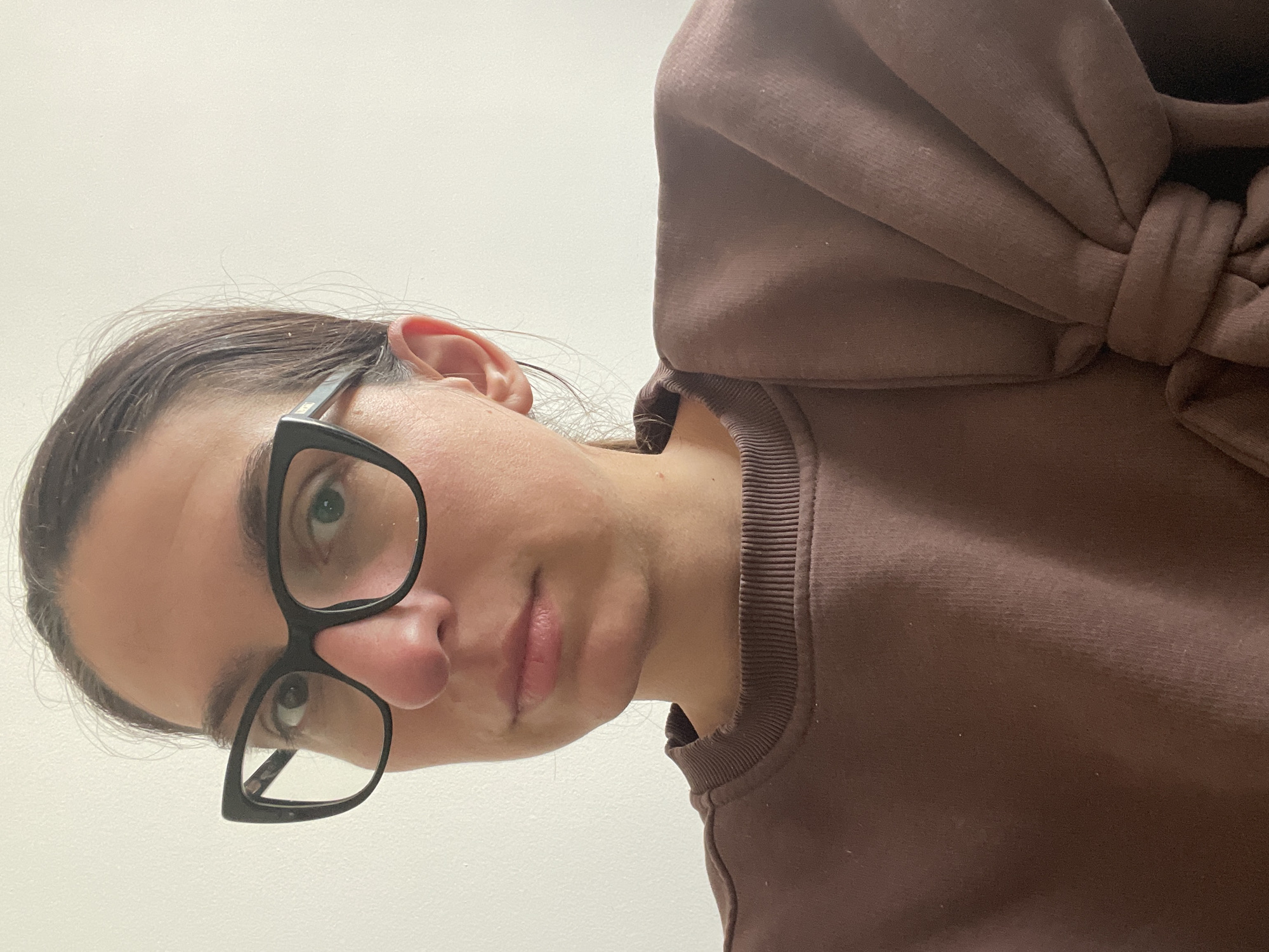 A woman with dark hair pulled back, wearing black-rimmed glasses and a brown sweatshirt with a decorative bow on the shoulder, is looking down at the camera. The background appears to be a plain, light-colored wall. The image is angled from above the subject. The location in Sozopol, Bulgaria is undetermined.
