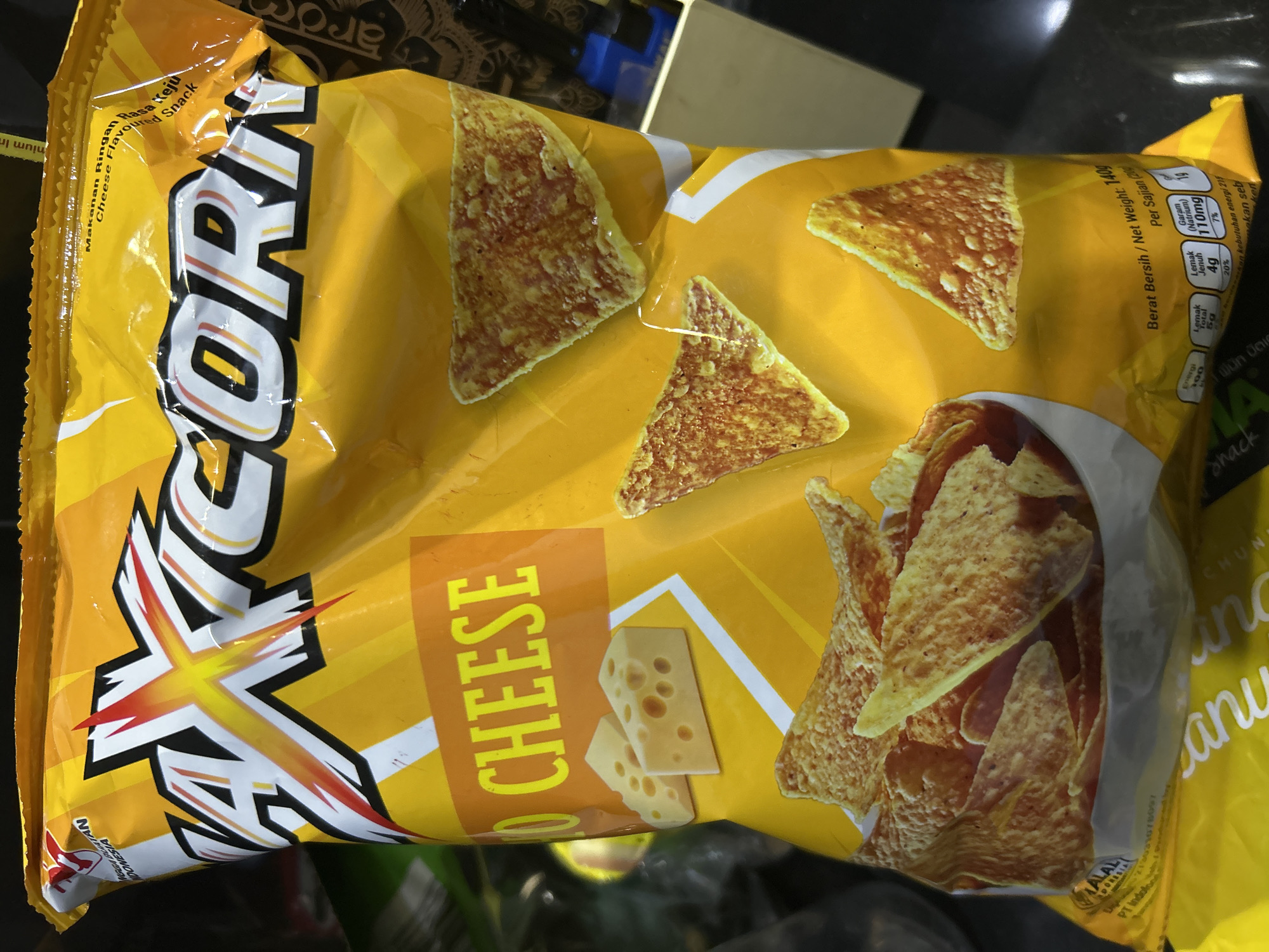 This is a close-up, eye-level shot of a brightly colored, yellow snack bag. The bag features the prominent brand name "AXICORN" in large, bold letters. The bag is designed with a vibrant yellow background and depicts several triangular-shaped chips, presumably the product inside, scattered across the front. There are also images of cheese chunks, reinforcing the flavor profile.
The text on the bag indicates that the product is "Makanan Ringan Cheese Flavour" which translates to "Cheese Flavored Snack" in English. Nutritional information is also visible, including "Berat Bersih / Net Weight: 140g" (Net Weight: 140g) and serving details for "Energi" (Energy), "Lemak Total" (Total Fat), "Lemak Jenuh" (Saturated Fat), and "Garam (Natrium)" (Salt (Sodium)). Some text also suggests the product is made in Indonesia.
The lighting suggests an indoor setting, possibly a store or a home, with the flash from the camera creating some glare on the glossy surface of the bag. The background is dark and slightly out of focus, indicating that the snack bag is the primary subject. There are no people visible in the image. The overall impression is a product shot designed to showcase the snack and its flavor.