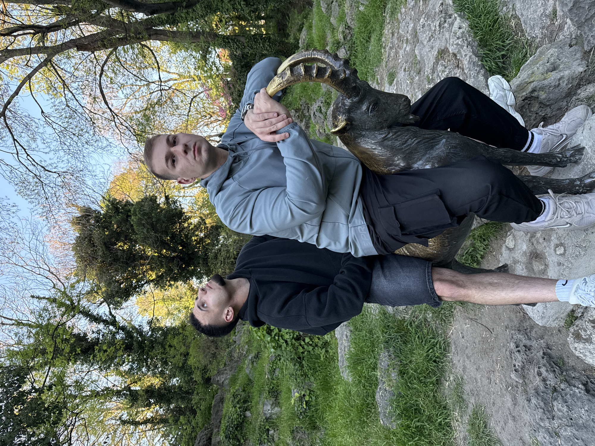 Two men are posing with a bronze statue of a goat in a park-like setting. The man on top, facing forward, wears a light gray hoodie and black pants. He is holding the goat's horns. The man below, facing upward, wears a black hoodie and dark gray shorts. Both men are wearing athletic shoes. The background includes trees, sky, grass, and rocks. The location appears to be in the city of Varna, Bulgaria.