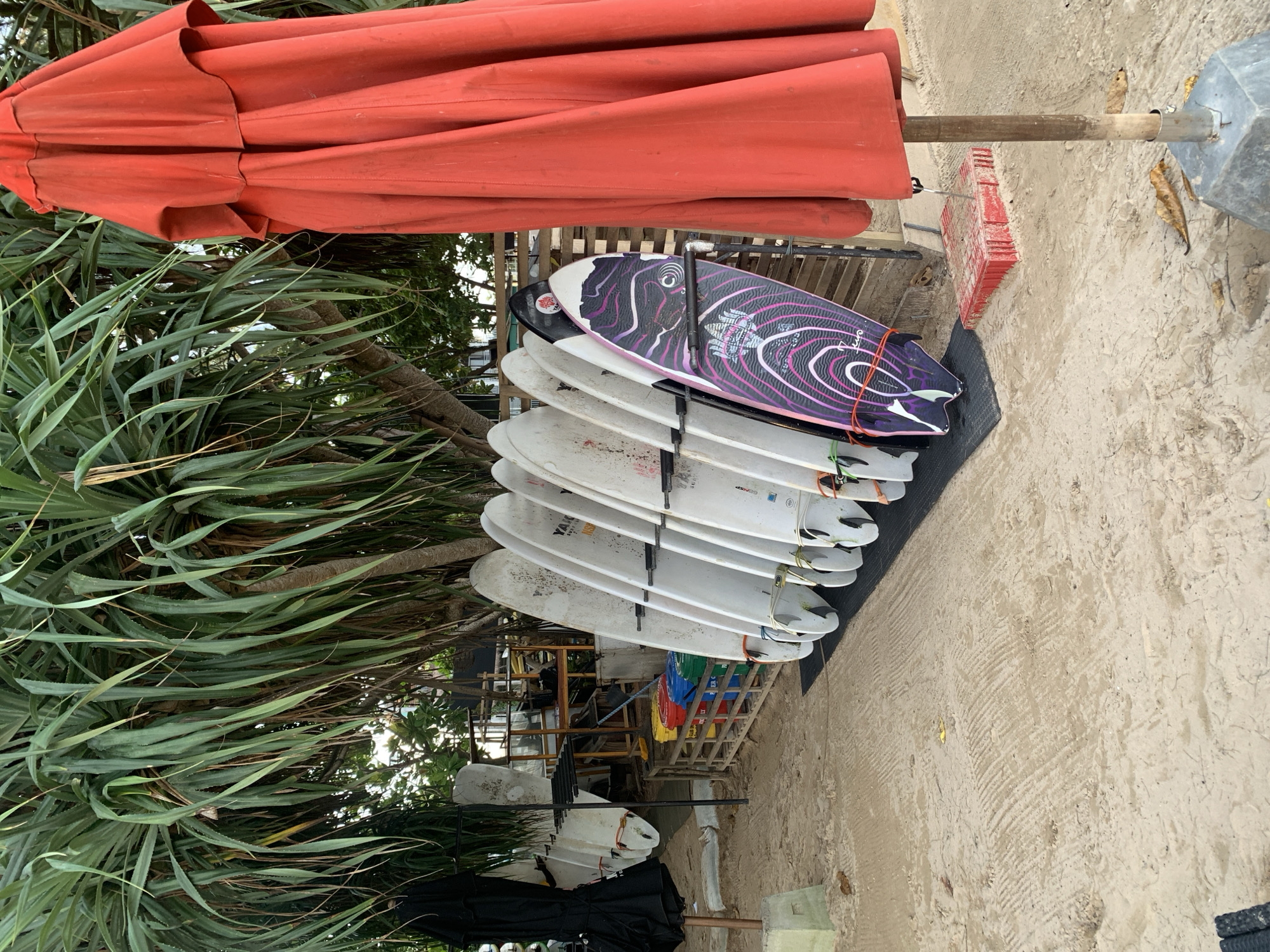 This photo was taken in Weligama, Sri Lanka. A stack of surfboards in a rack are positioned beneath a large, closed, red umbrella. Next to the surfboards is a leafy tree. The ground is sandy. There are no people.