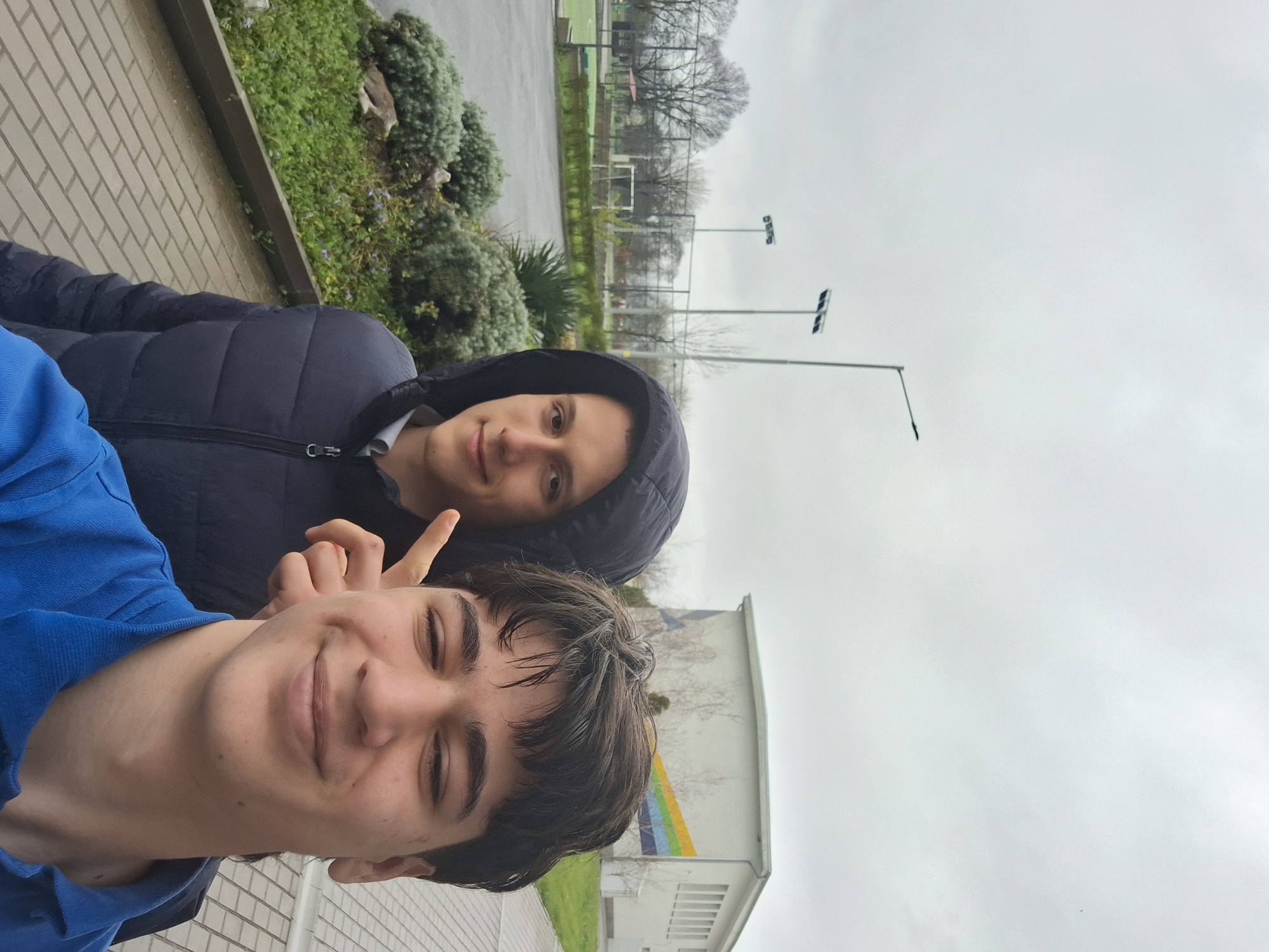 Two young men are in a selfie-style photograph, likely outdoors in Burgas, Bulgaria. The foreground figure is in a blue shirt, smiling with closed eyes and dark, wet hair. Behind him and slightly to the right is another young man wearing a hooded, dark blue jacket, also looking at the camera. The background features a paved walkway, greenery, a building with a rainbow-colored design, light poles, a fenced-in area, and an overcast sky.