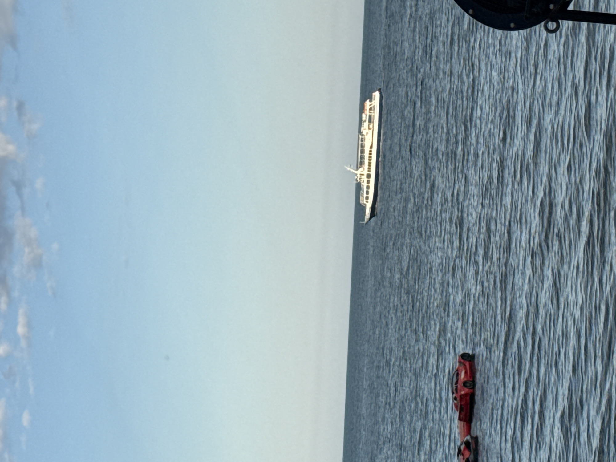 The media file displays a scene at sea under a clear, light blue sky with scattered clouds. A long, white passenger ferry with multiple decks sails on the deep blue water. In the foreground, near the right edge of the frame, two red buoys are visible floating on the water's surface. The image appears to be taken during the daytime, likely late afternoon or early evening, given the soft lighting. The sea is calm with gentle ripples. The composition suggests a view from another vessel or a waterfront structure. No people are clearly discernible in the image. The scene evokes a sense of peaceful travel or maritime activity.