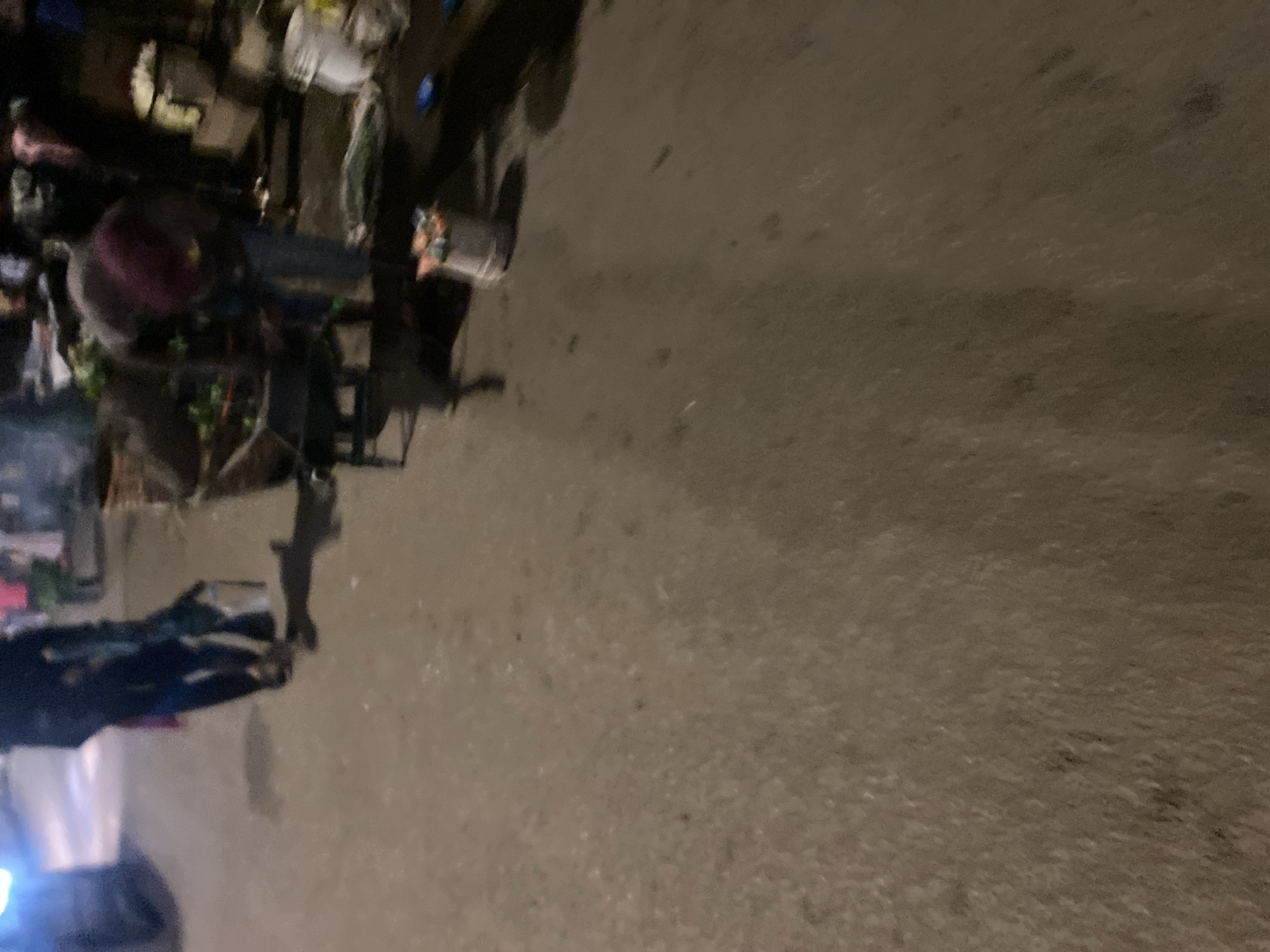 The image, taken at what appears to be night or late evening due to the dim lighting, depicts an outdoor scene on a rough, dark grey-brown ground, likely a street or market area.
The main subject is a stationary motorcycle or scooter, heavily laden with various items, suggesting it is used for transporting goods or as a mobile vending setup. On the left side of the vehicle, multiple containers and baskets are visible, including a prominent reddish-purple bowl, possibly holding produce or merchandise. The background on the left is dark and indistinct, with vague shapes that could be other vehicles or structures. A bright light source, possibly a vehicle's headlight or a lamp, is visible in the lower-left section of the image (when oriented correctly).
No identifiable people are clearly visible, and there is no discernible dynamic activity taking place; the scene is static. The overall impression is that of a practical, perhaps commercially active, roadside or market environment. No visible text can be discerned in the image.