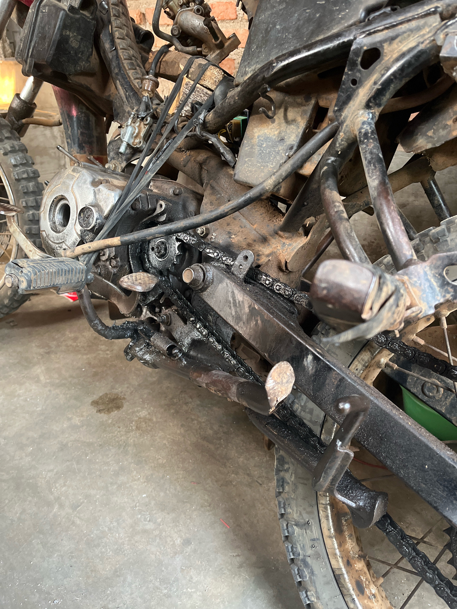 The image depicts a close-up, detailed view of the engine and drivetrain of a heavily used and possibly damaged motorcycle. The scene is set on a concrete floor, with what appears to be a brick wall in the background. This location is identified as being in the city of Kebumen, Indonesia.
The motorcycle itself is in a state of disrepair or extensive use. The engine casing is visible, showing some wear and tear, with oily residues coating parts of it. A prominent chain runs through the drivetrain, also visibly coated in oil or grease. Various metal components, including the frame, footpegs, and parts of the suspension or exhaust system, are present. These components exhibit signs of rust and significant grime, suggesting prolonged exposure to the elements and heavy use.
Several wires and cables are routed throughout the engine area, some appearing frayed or improperly secured. A section of the rear tire is visible, further indicating the motorcycle's operational status. The overall impression is one of a functional but neglected piece of machinery, likely in a workshop or storage area within Kebumen. There are no people or direct actions being performed on the motorcycle in this specific frame. The environment suggests a practical, perhaps informal, setting for the maintenance or storage of this vehicle.