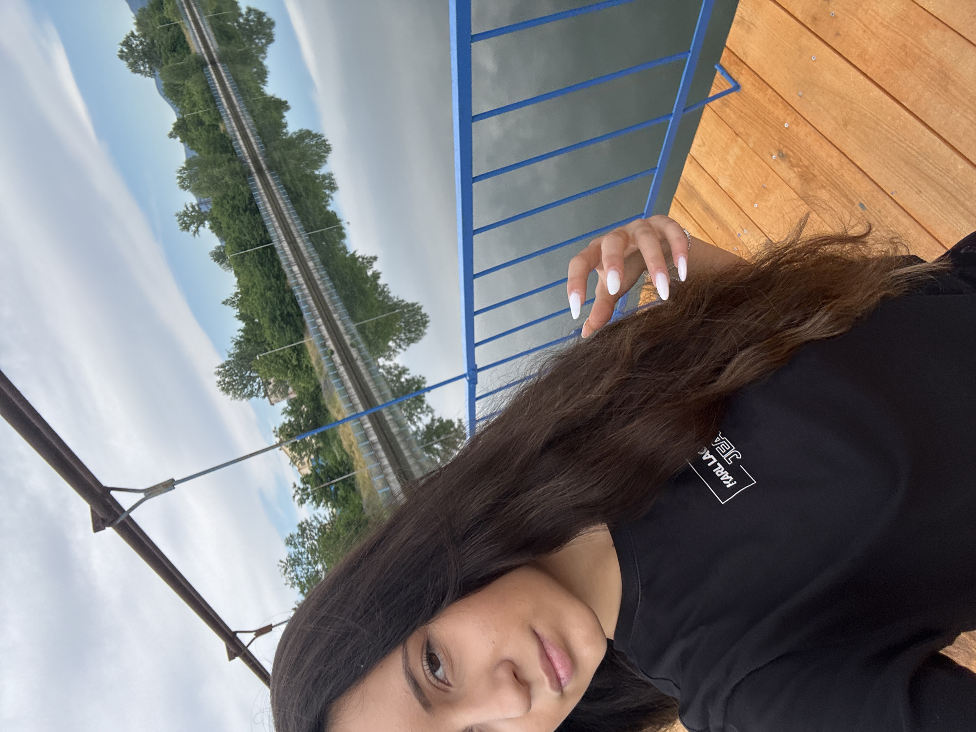 A young woman with long, dark hair takes a selfie on a wooden pier with a blue metal railing. She is wearing a black t-shirt with a white logo that reads "KARL LAGERFELD JEANS." Her nails are painted white. Behind her, a body of water reflects the sky and trees. The location is unknown, but appears to be an outdoor setting by a lake or river, possibly in a park or recreational area. The overall impression is casual and relaxed.