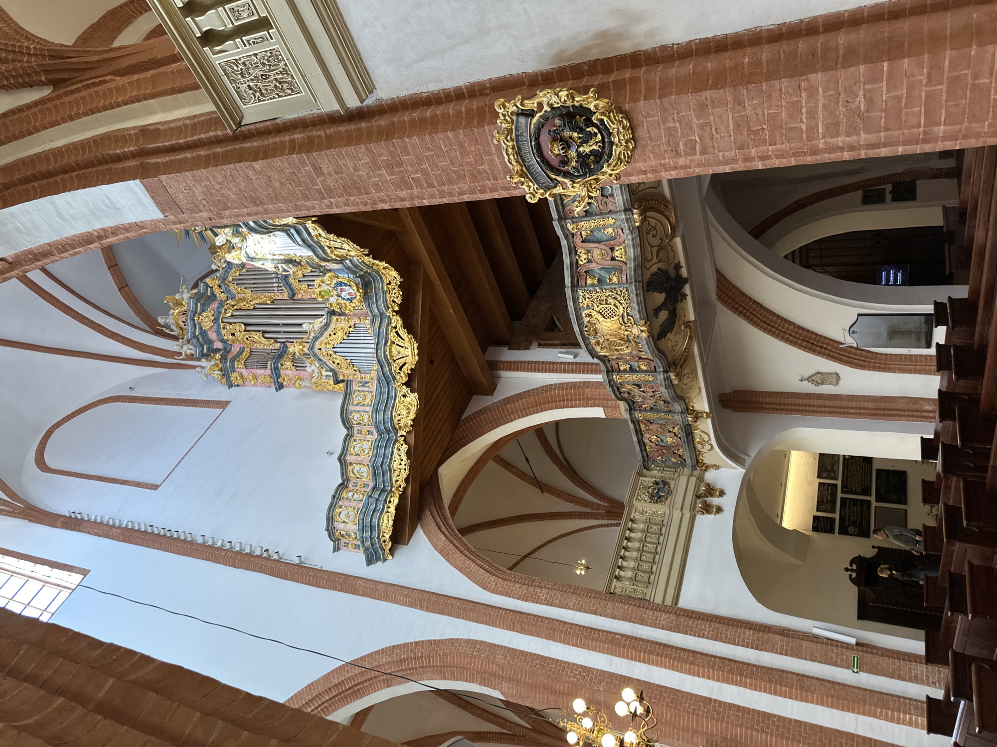 The image depicts the interior of a church, likely located in Wrocław, Poland, given the architectural style. A prominent pipe organ, adorned with gold and blue decorative elements, is mounted on the wall. The interior features a combination of red brick and white plaster surfaces. Arches are a recurring structural element. Decorative carvings and crests are attached to the brick beams. A chandelier is visible in the lower part of the image. In the background, through an archway, some people can be seen. Church pews line the floor to the right.