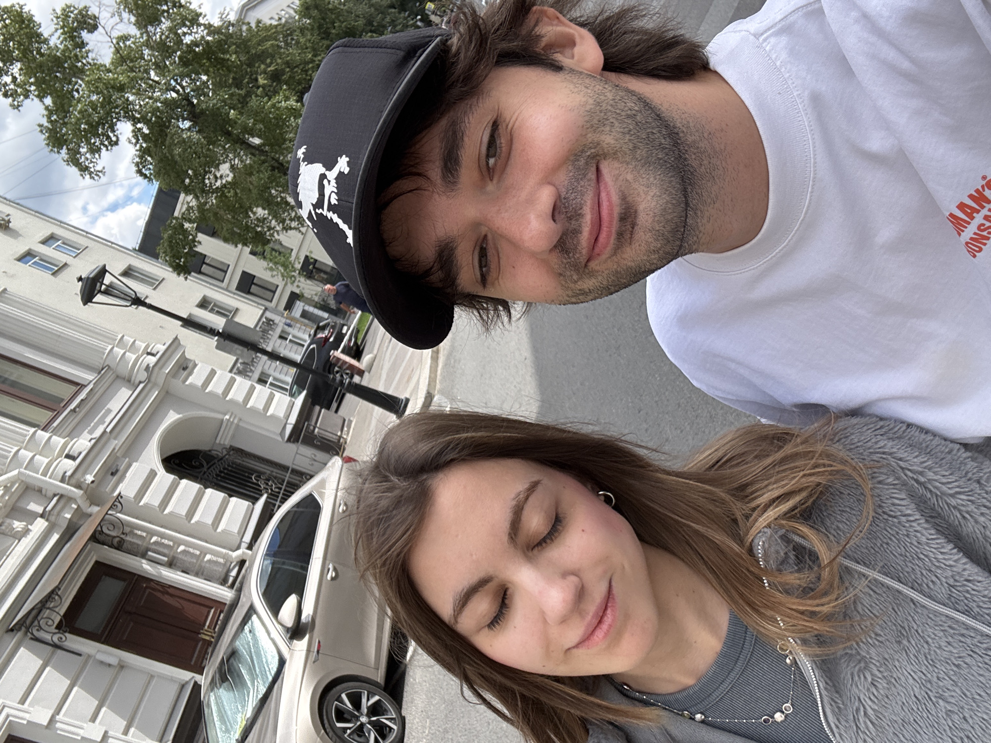 The media file is a selfie taken by two people outdoors, likely in a city. The person in the foreground on the right is a woman with long, light brown hair, closed eyes, and a gentle smile, wearing a grey fuzzy jacket over a grey shirt and a silver necklace. The person in the background on the left is a man with dark hair, wearing a black baseball cap with a white graphic, and a white t-shirt with red text that appears to read "MAN'S CONS..." and "...ONSAL". He is looking towards the camera with a slight smile.
The scene behind them includes a light-colored building with ornate architectural details, a dark-colored car parked on the street, a lamppost, and a large tree with green leaves. The sky above is partly cloudy. The overall impression is a casual, daytime snapshot. The presence of the building and the car suggests an urban setting, and the provided location context confirms it is Moscow, Russia. The time of day appears to be daylight, and the weather seems pleasant. The subjects appear relaxed and happy.