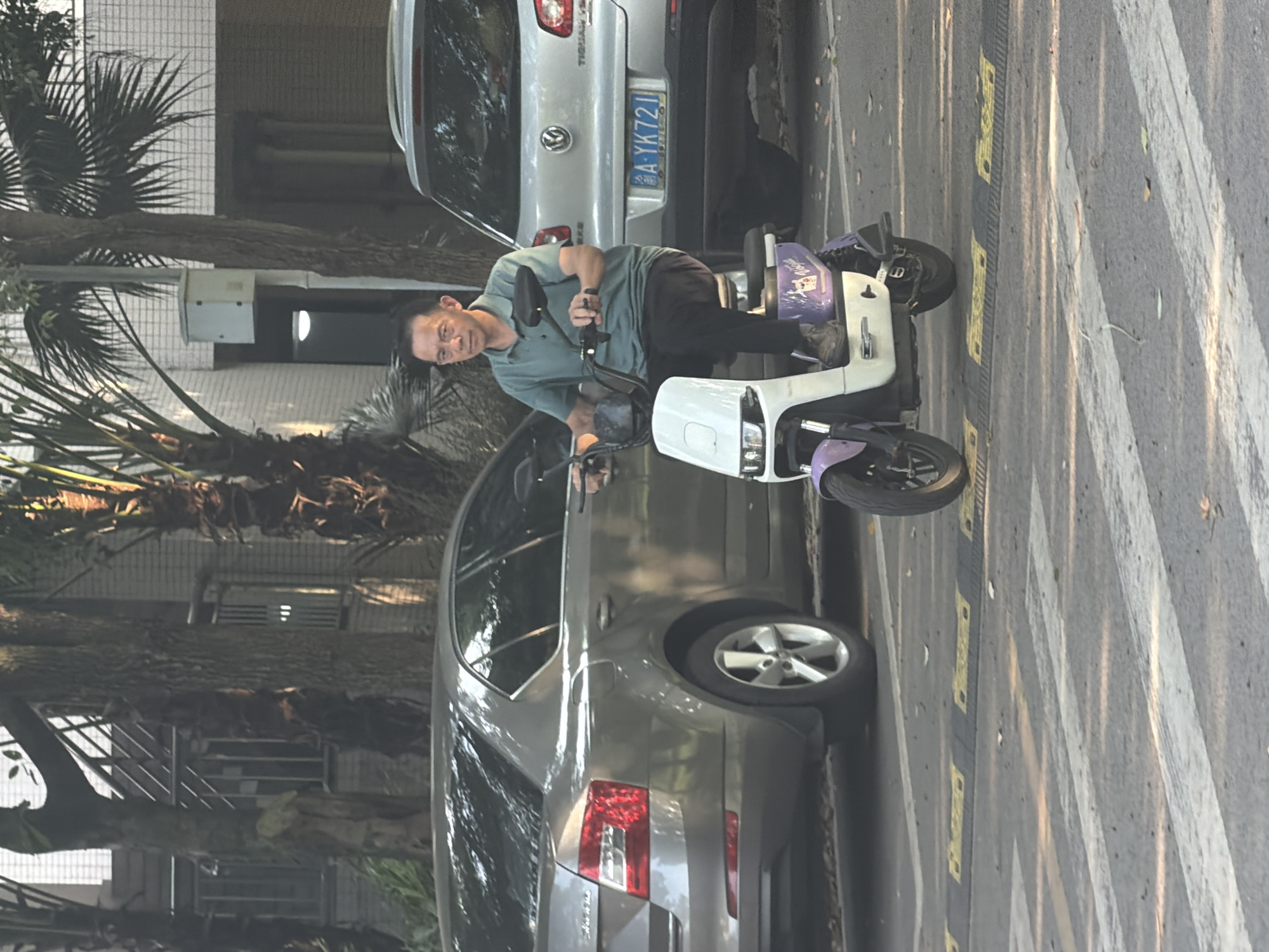 A middle-aged East Asian man is riding a white and purple electric scooter across a pedestrian crossing. He is wearing a green polo shirt and dark pants. He is looking forward. The scooter has the word "Vogue" written on its side. Behind him, there is a silver Volkswagen Tiguan SUV with a license plate that reads "A YK721." To the left of the SUV, there is a silver sedan. In the background, there are buildings and palm trees. The scene appears to be a street in Jiangjin, China.