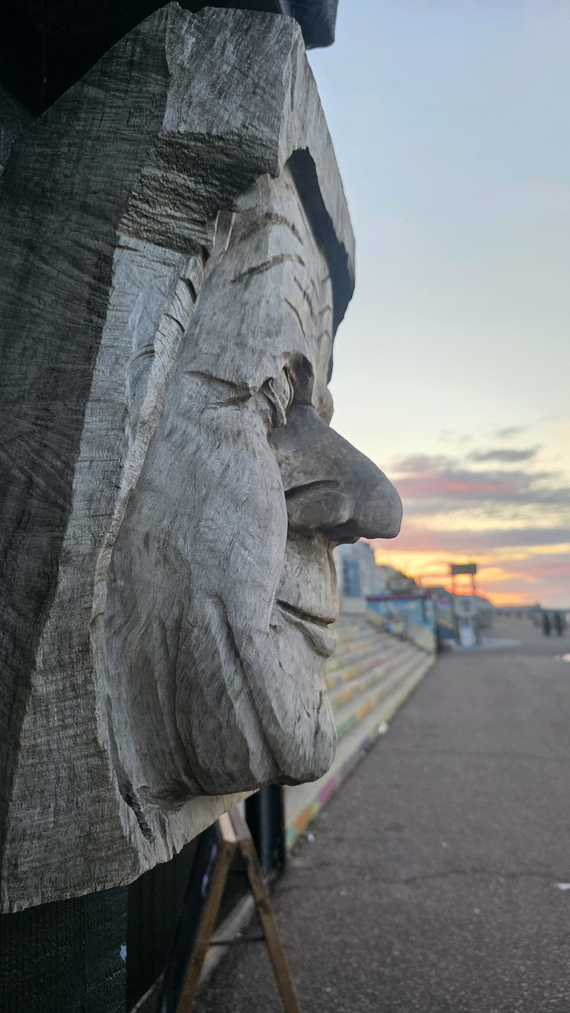 A close-up, side profile of a weathered, carved wooden face is the focal point of this image, positioned on the left side. The carving depicts a prominent nose, a heavy brow, and a deeply etched mouth. The texture of the wood is rough and grooved, showing signs of age and exposure to the elements. Behind the carving, the background opens up to reveal an outdoor scene at dusk. A paved pathway stretches into the distance, flanked by a concrete staircase on the left, which is adorned with colorful graffiti. The sky above is a gradient of pale blue, transitioning to hues of orange and pink as the sun sets, with scattered clouds. In the distance, silhouetted figures can be seen walking along the pathway, and a structure, possibly part of a pier or boardwalk, is visible on the horizon. The location is identified as St Leonards, United Kingdom, suggesting an urban coastal environment.