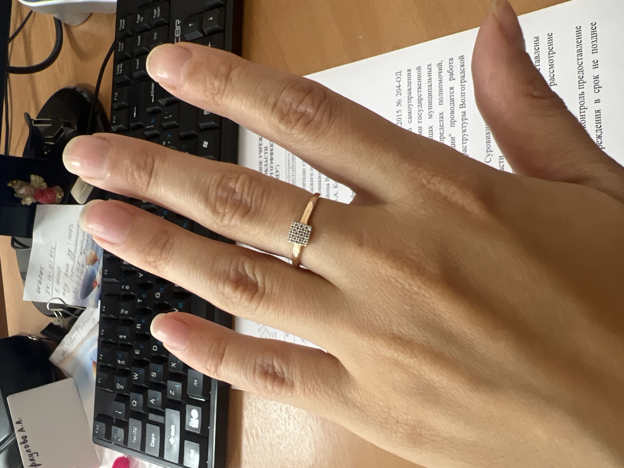The image shows a close-up of a person's hand with a ring on the ring finger, positioned over a document and a keyboard. The document appears to be an official paper with Russian text, mentioning "Волгоградской" which translates to "Volgograd", suggesting the location is in Volgograd, Russia. The keyboard is a standard QWERTY layout, visible in the background. There are also other papers and office supplies on the desk. The hand is lightly tanned with clean, manicured nails. The ring is delicate with a small, square, diamond-studded design. The lighting suggests an indoor office environment.