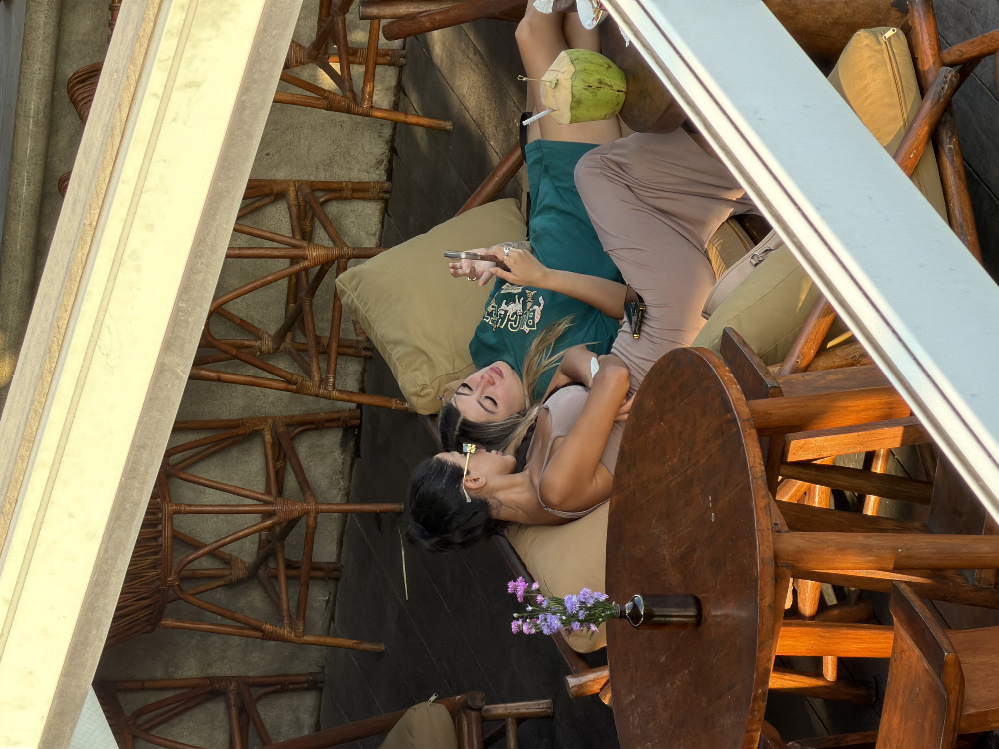Two young women are relaxing on a cushioned bench on a wooden deck, likely at a beachside establishment in Mengwi, Indonesia. The woman on the left, with dark hair pulled back, is wearing a light-colored tank top and a light brown maxi skirt. She is looking down, possibly at her phone or something on the table. The woman on the right, with blonde hair, is wearing a teal t-shirt and is lying on her side, looking at her phone which she holds in her hand. A green coconut with a straw is on the table next to her. A wooden table with a small vase of purple flowers sits in the foreground. In the background, there are several tall, wicker-backed stools and a wooden railing. The overall scene suggests a casual, leisurely atmosphere.