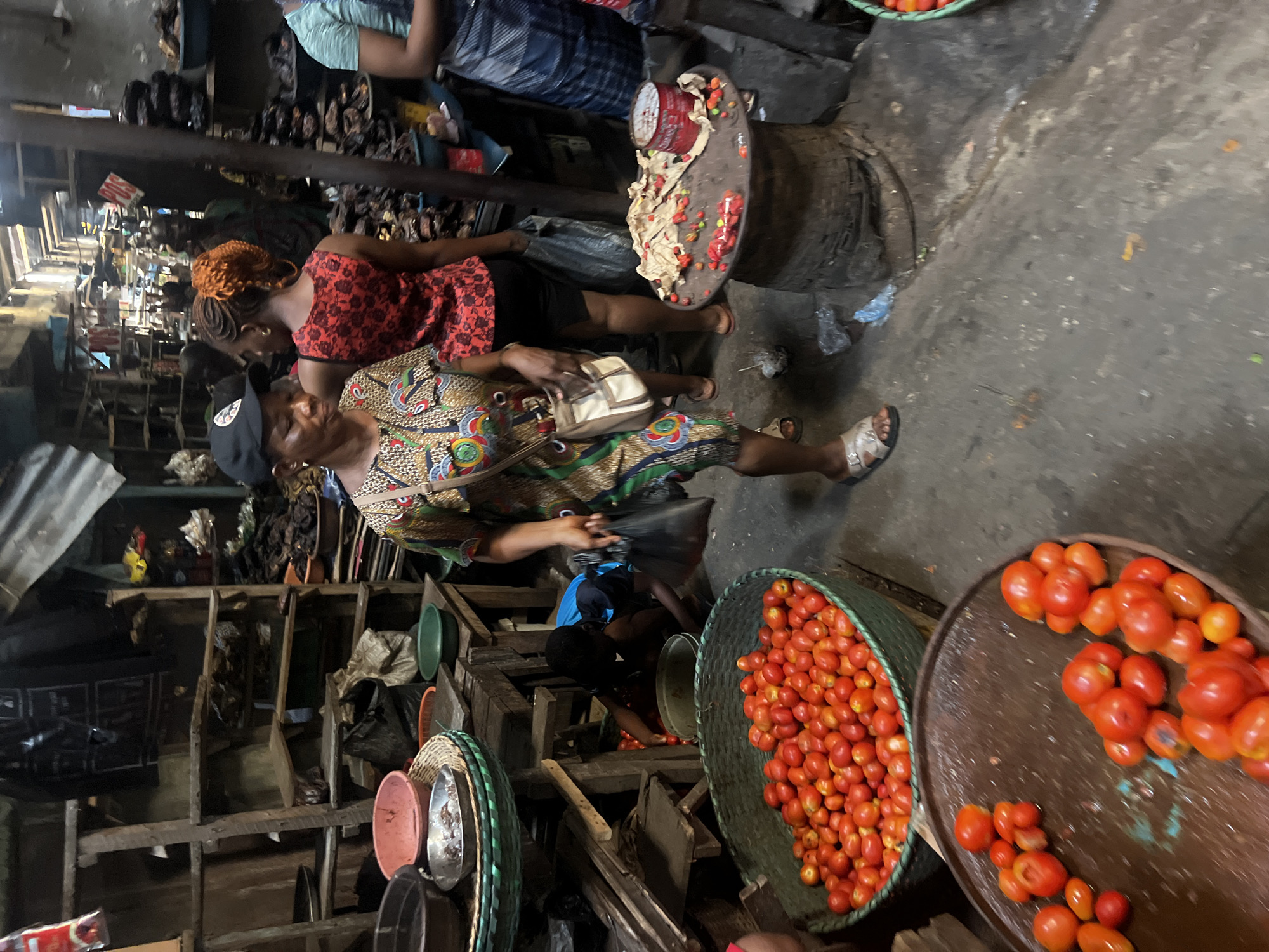 A photograph shows the inside of a market in Nkpor, Nigeria. Two women are looking at or buying red tomatoes. One woman is wearing a red floral top and the other is wearing a colorful dress and a black cap. One woman is holding a white purse. One is standing near a wooden stool with a red bucket on top. There are also baskets full of tomatoes on the ground. The market stalls are made of wood.