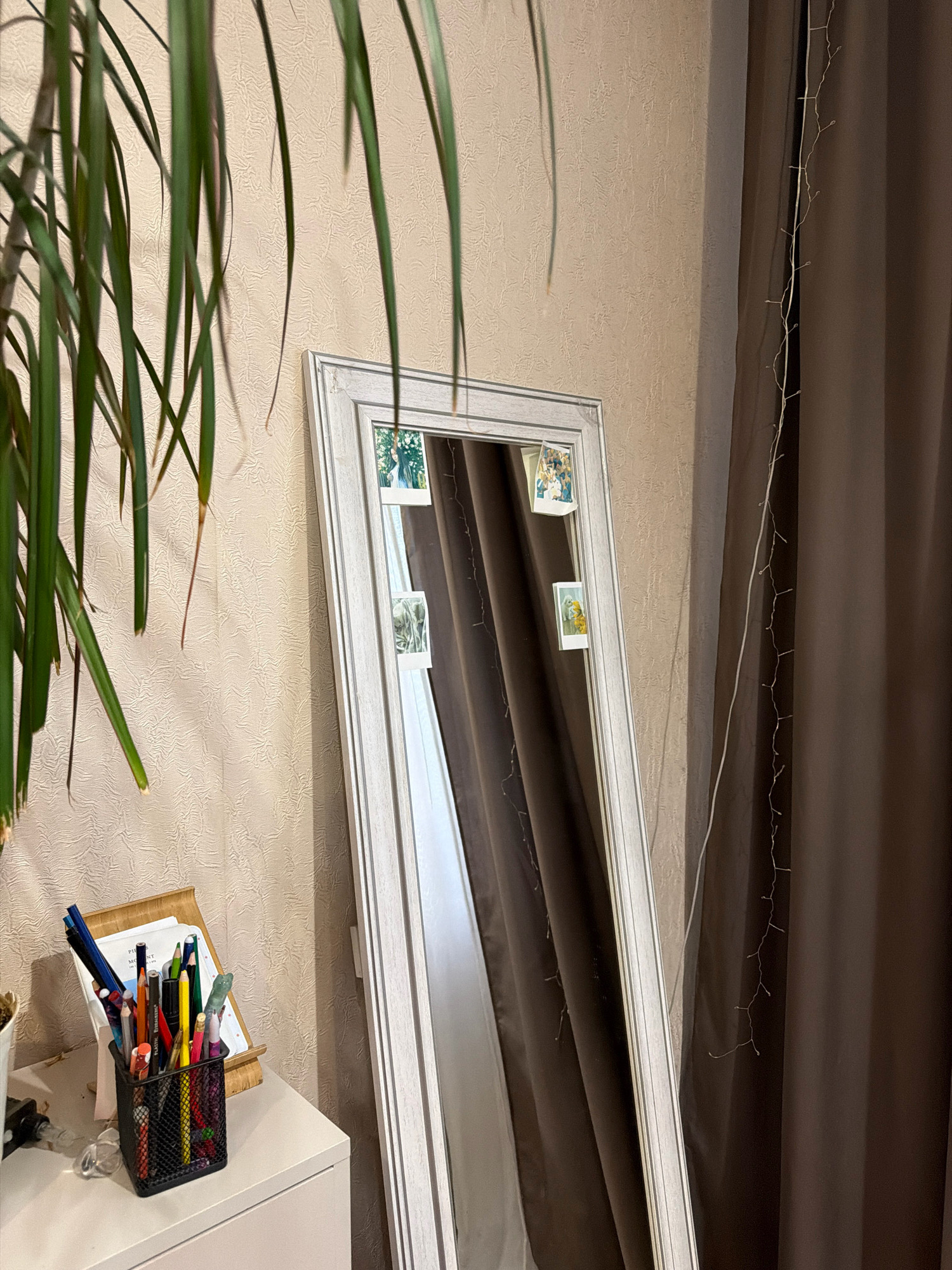A full-length mirror with a white frame stands leaning against a beige, textured wall. The mirror reflects a portion of the room, including a dark brown curtain and a string of small, white lights draped vertically. Attached to the mirror's frame are three small, square photographs. To the left of the mirror, a white cabinet is visible, topped with a black mesh organizer filled with pens and pencils. Behind the organizer is a wooden easel holding a few papers. In the upper left corner of the image, the long, green leaves of a plant hang down, partially obscuring the scene. The lighting in the image is soft, suggesting it was taken indoors during the day.