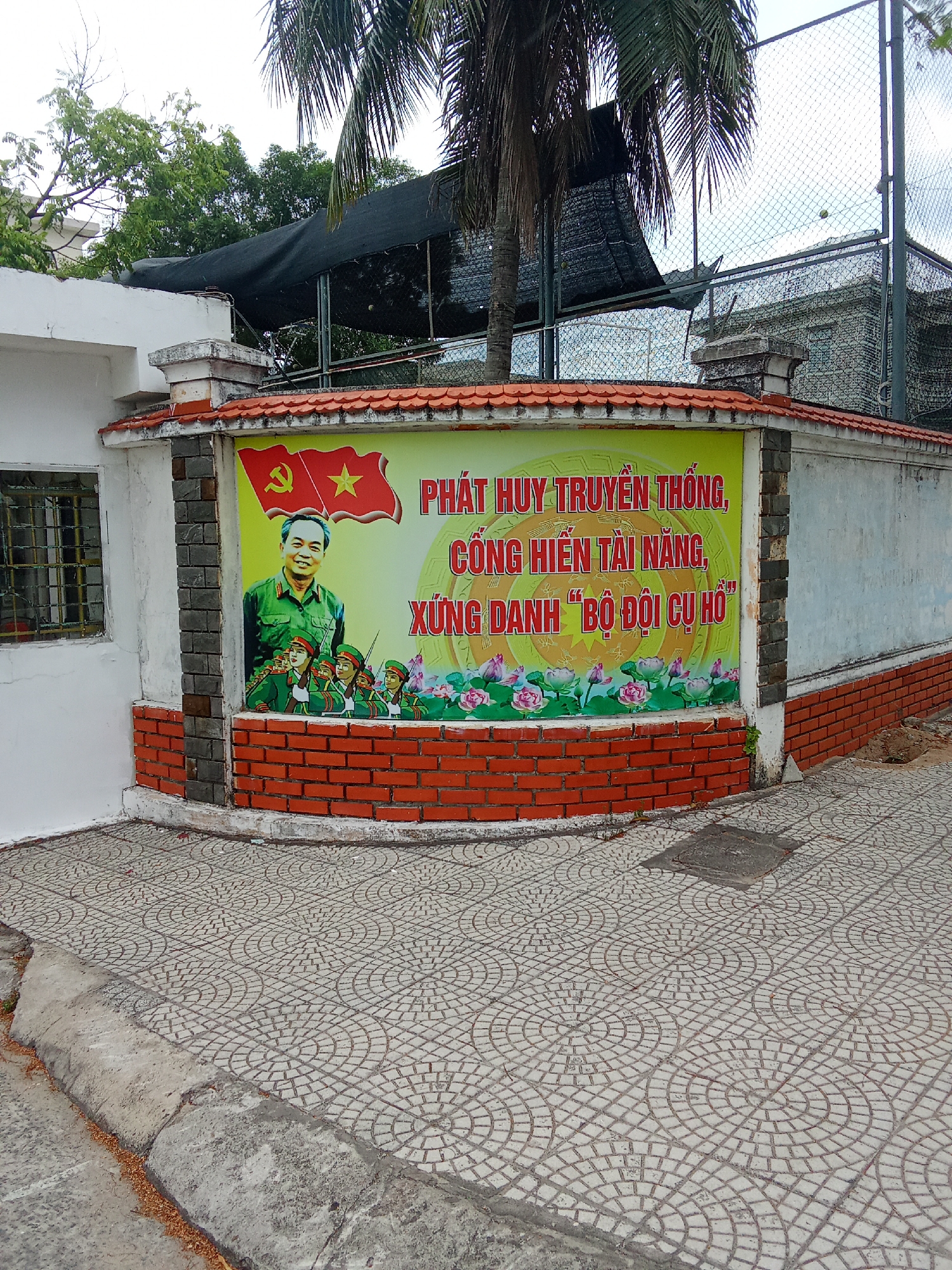 The image shows a mural painted on a wall in the city of Đà Nẵng, Vietnam. The mural features a portrait of a man in a military uniform, a Vietnamese flag, and several figures in military uniforms marching. The text on the mural is in Vietnamese and appears to be a patriotic slogan. The mural is surrounded by a brick border and is located next to a white building with a barred window. In the background, there are trees and a chain-link fence. The ground in front of the mural is paved with square tiles.