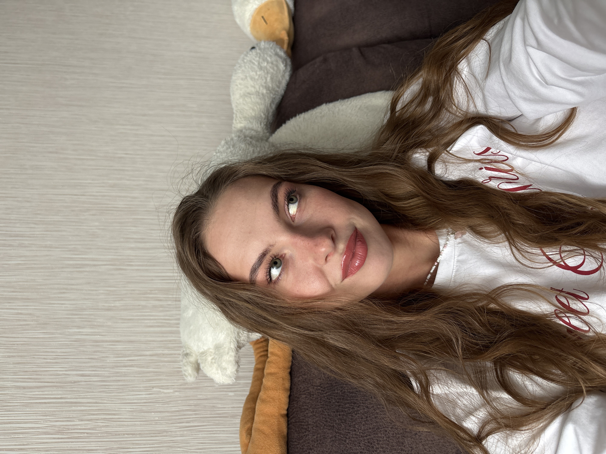 The image features a young female individual in an indoor setting. Visual information within the frame does not confirm the specific location as Perm, Russia. The woman, appearing to be in her twenties, is captured in a close-up, chest-up shot. She is looking to her left (viewer's right) with a soft, upward gaze and a subtle smile. She has long, wavy, light brown hair, green eyes, and light skin with visible freckles on her nose and cheeks. Her makeup includes winged eyeliner, pink eyeshadow, and a glossy, berry-toned lipstick. She is wearing a white long-sleeved sweatshirt with partially visible red embroidered text, and a white beaded necklace with a small pink charm is around her neck. She is seated or leaning against a dark brown couch. Behind her, resting on the couch, are several plush toys: an orange elongated object on the left, a white stuffed goose, and a white stuffed duck further to the right. The wall behind the couch is covered in light beige or off-white wallpaper featuring a fine, vertical ridged texture.