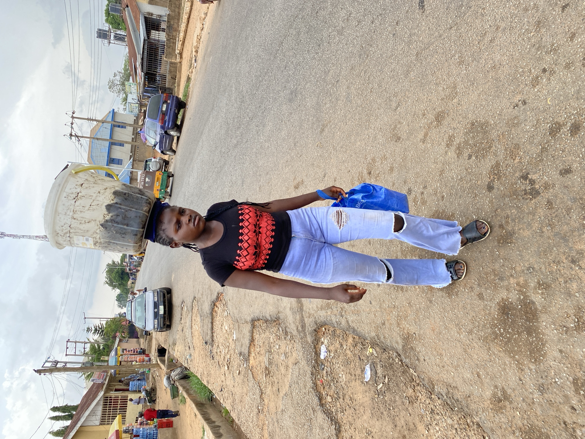 A young woman stands on a cracked asphalt road in Bukuru, Nigeria. She carries a large, dirty bucket on her head and a blue plastic bag in her left hand. She wears a black t-shirt with a red patterned design, light blue ripped jeans, and black sandals. The road is flanked by buildings and vehicles, including cars and a tricycle. A water vendor and several cases of bottled water are visible near a building on the side of the road. A tall telecommunications tower is in the background. The sky is overcast.